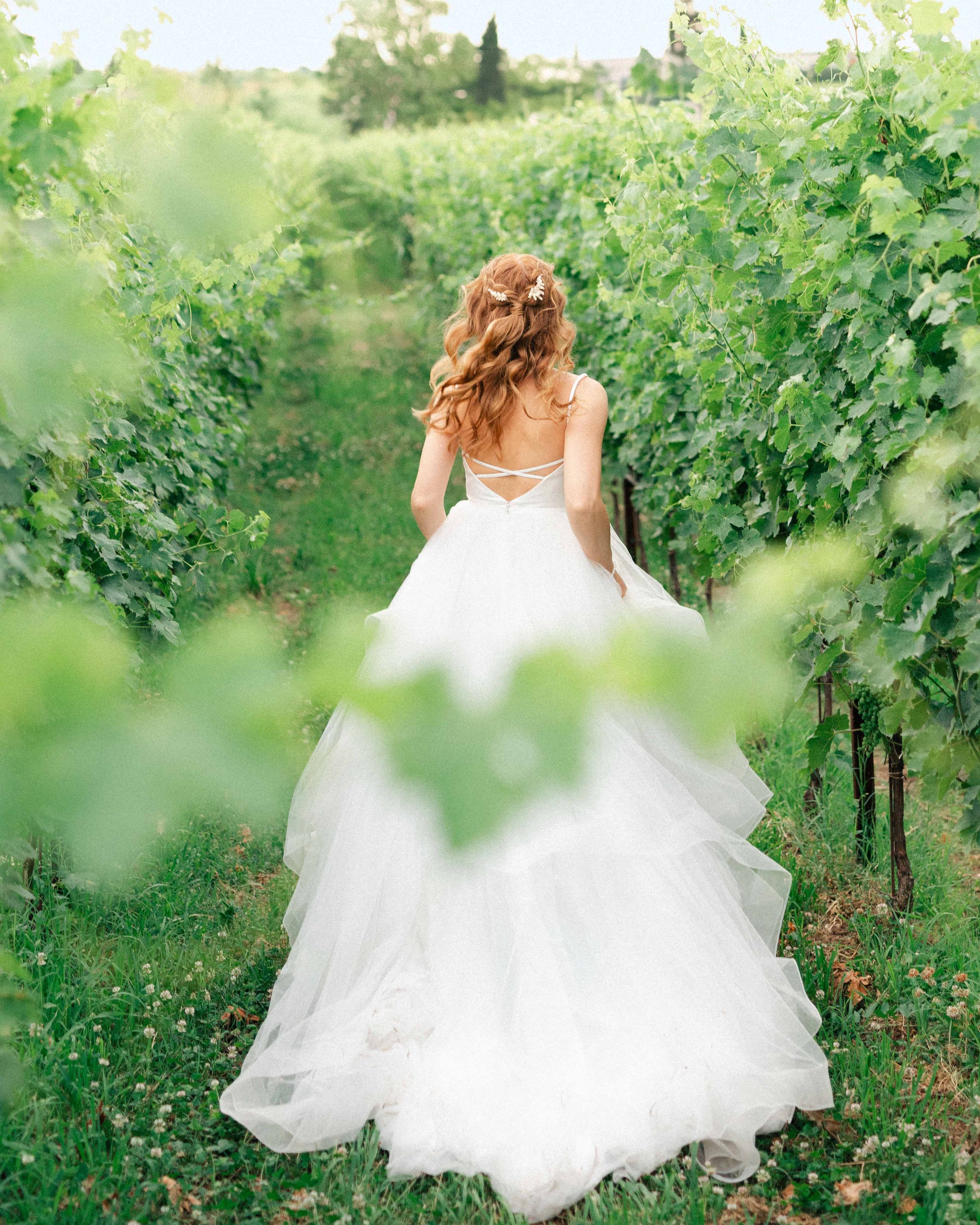 A woman in a white dress with a full skirt walking through a vineyard with green leaves and vines on either side.