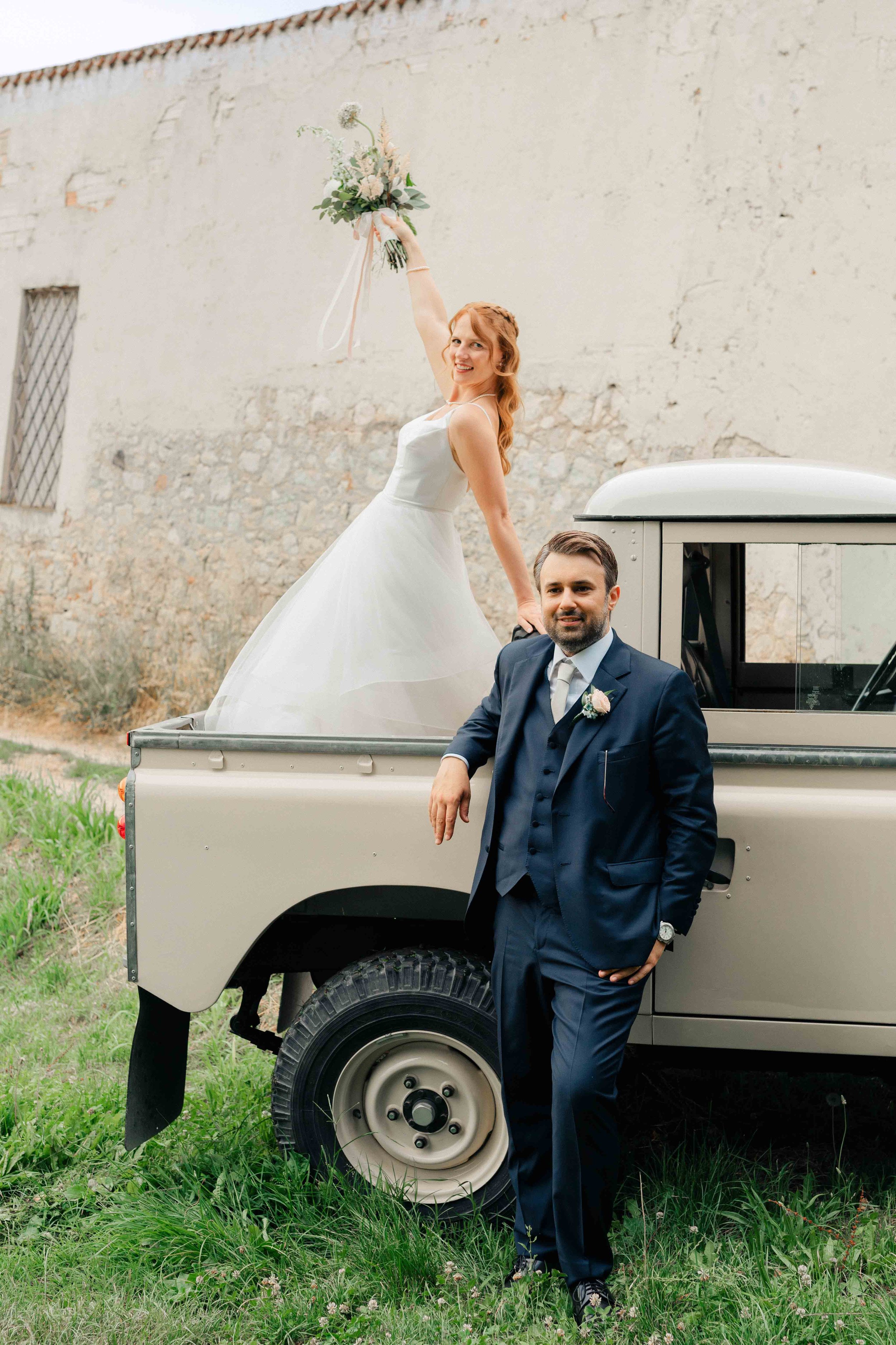 A woman in a wedding dress holding a bouquet, smiling, sitting on a vintage truck. A man in a dark suit standing next to the truck, with his hand in his pocket, smiling. The background features an old wall and grassy field.
