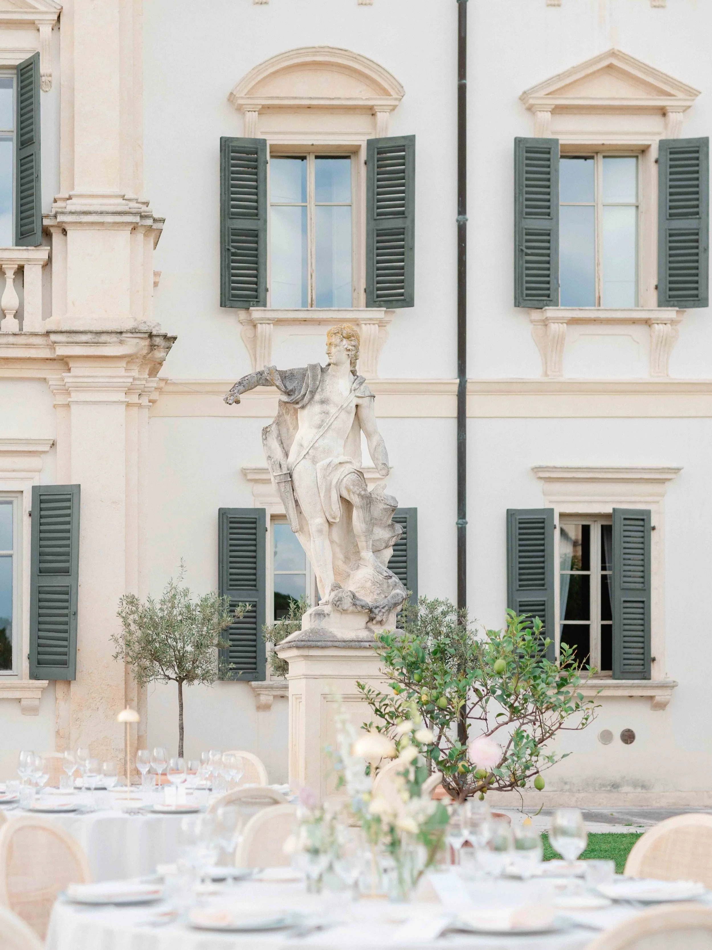 Outdoor dining setup with chairs and tables, a marble statue of a classical figure, and a building with windows and shutters in the background.