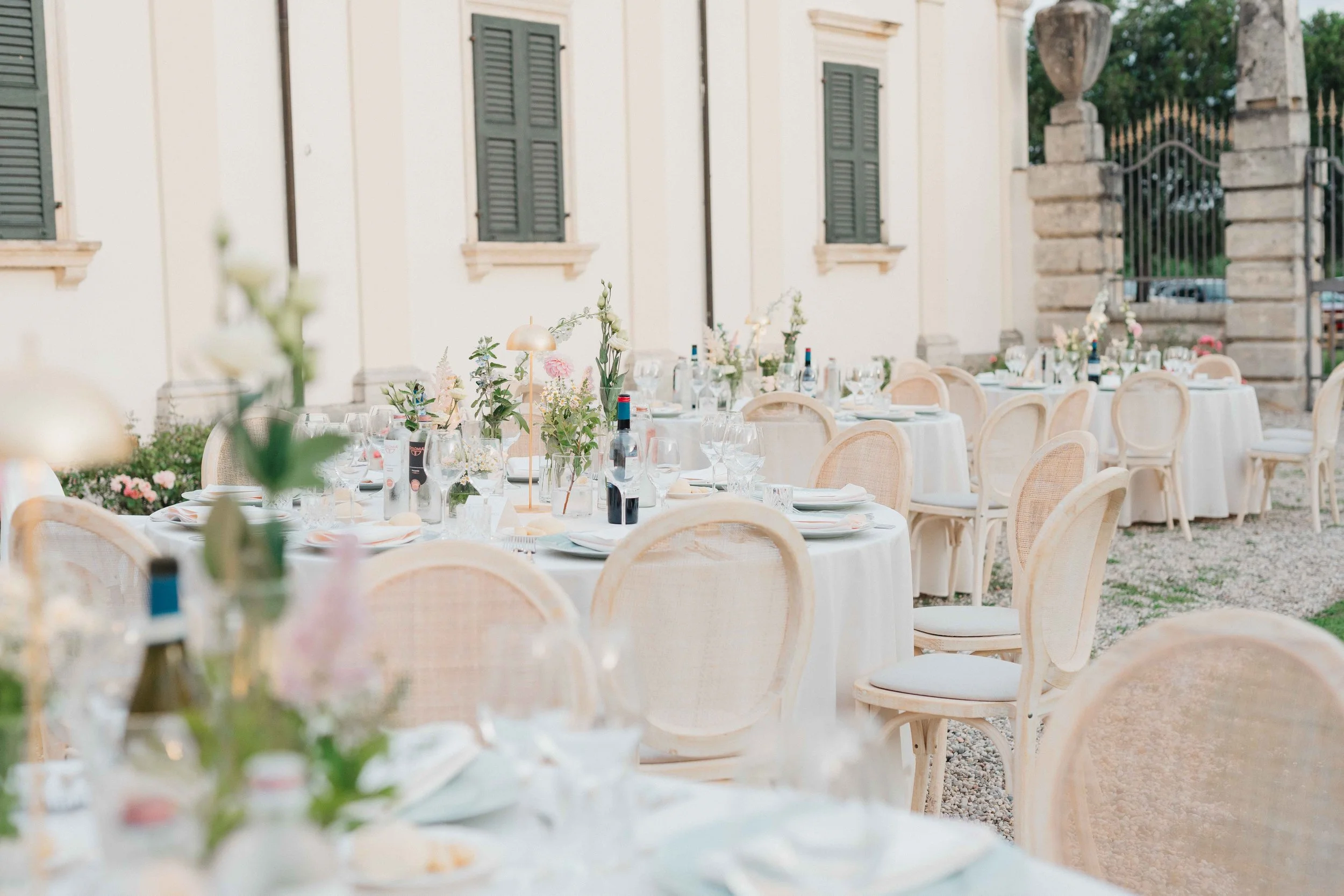 Outdoor banquet setup with round tables decorated with floral centerpieces, chairs with woven backs, and a white building with green shutters in the background.