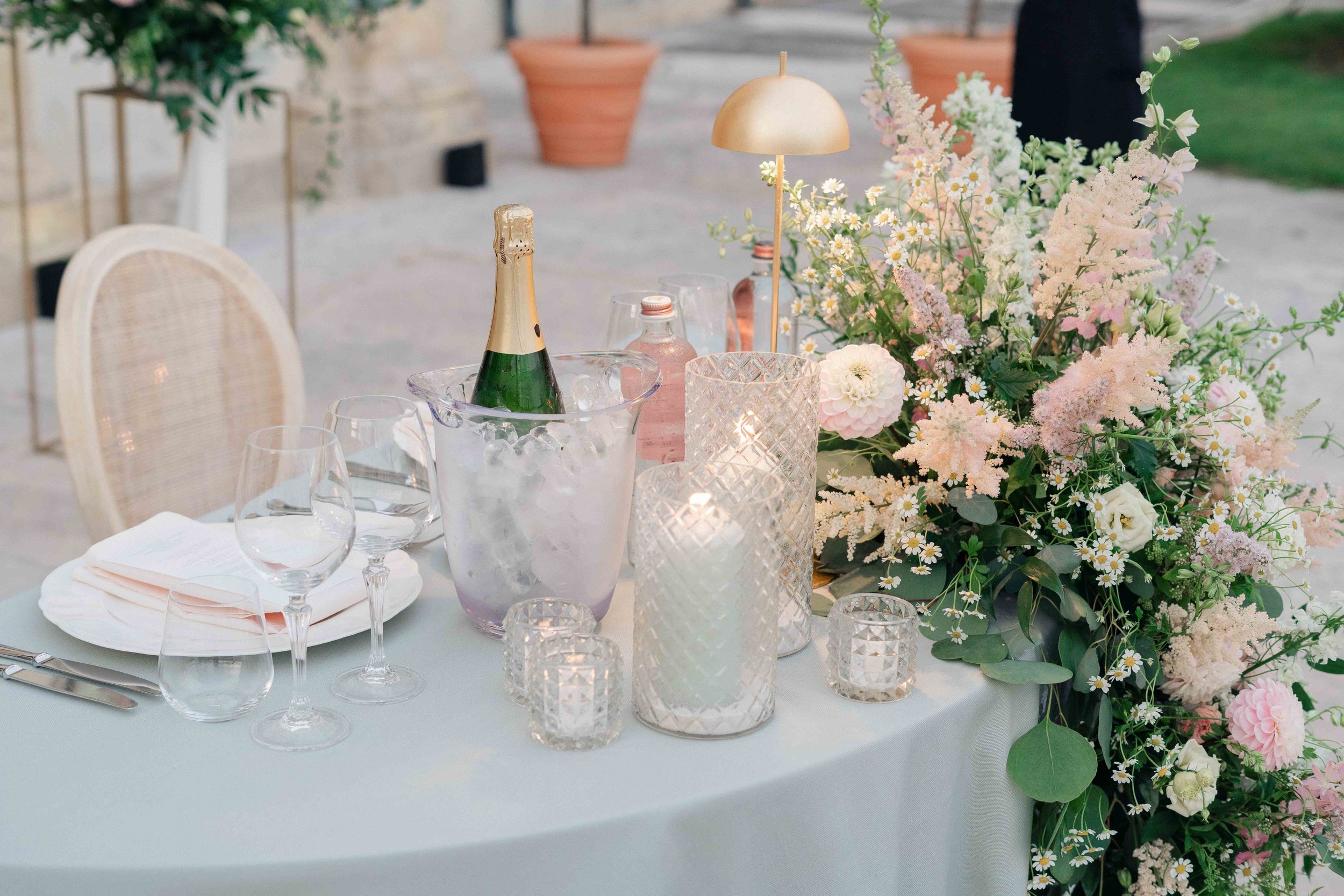 A table set for a celebration with a floral centerpiece, candles, and a champagne bottle in an outdoor setting.