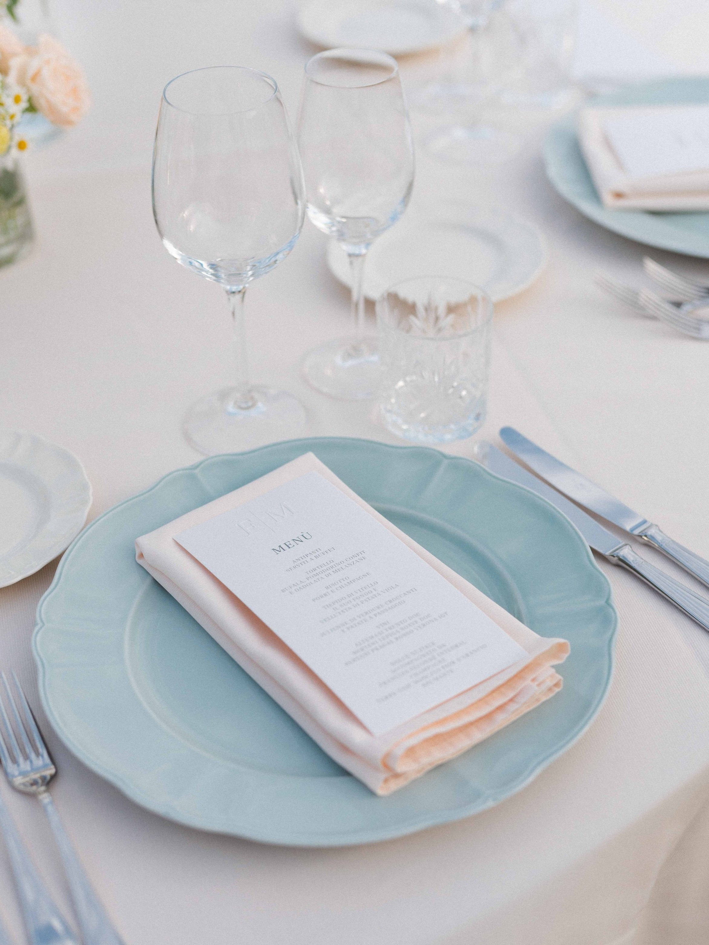 Elegant table setting with a blue plate, a folded printed menu, silver cutlery, and clear glassware, including two wine glasses, on a white tablecloth with a floral centerpiece visible in the background.