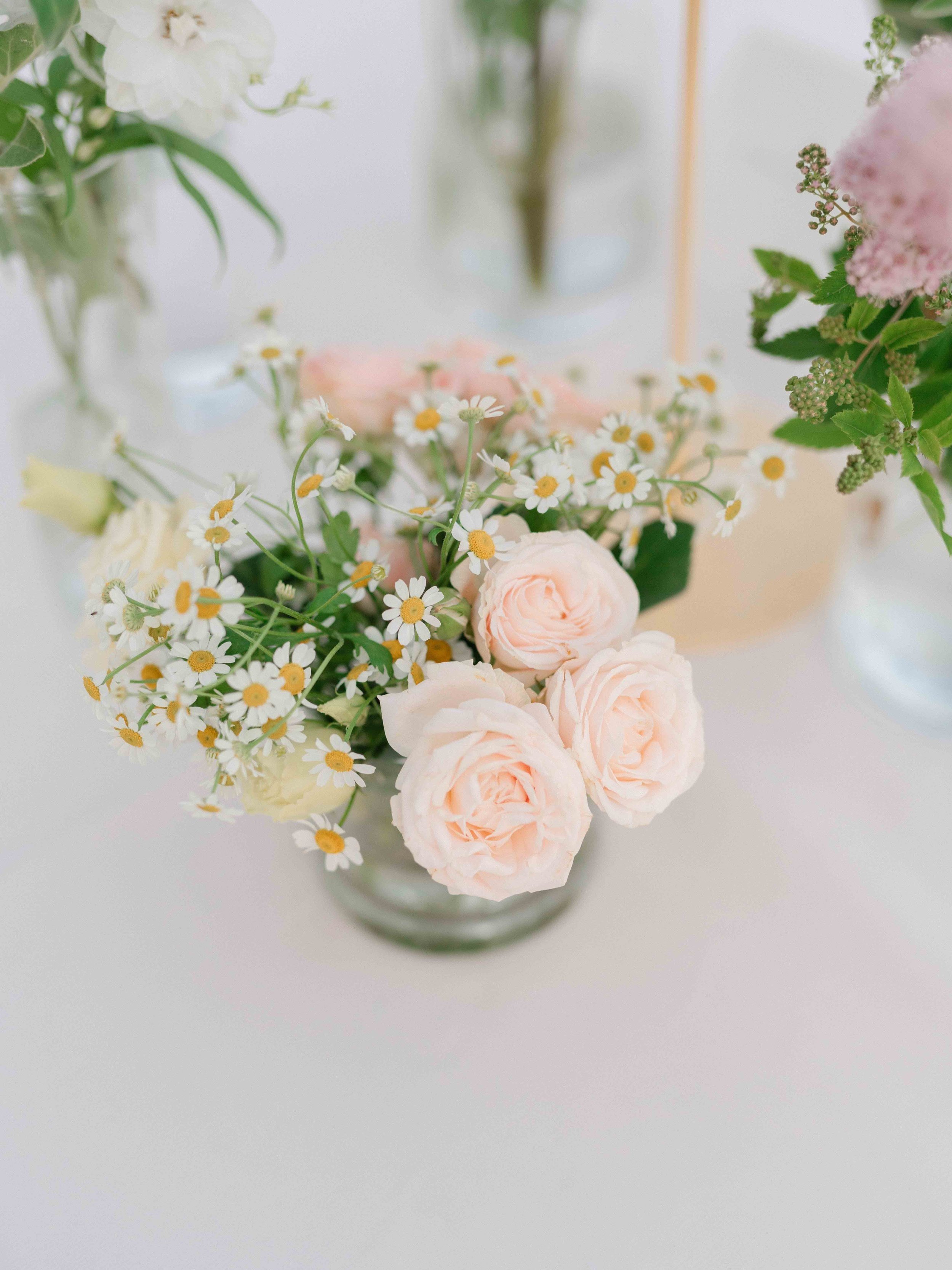 Arrangement of white daisies, pink roses, and greenery in a clear glass vase on a white surface.