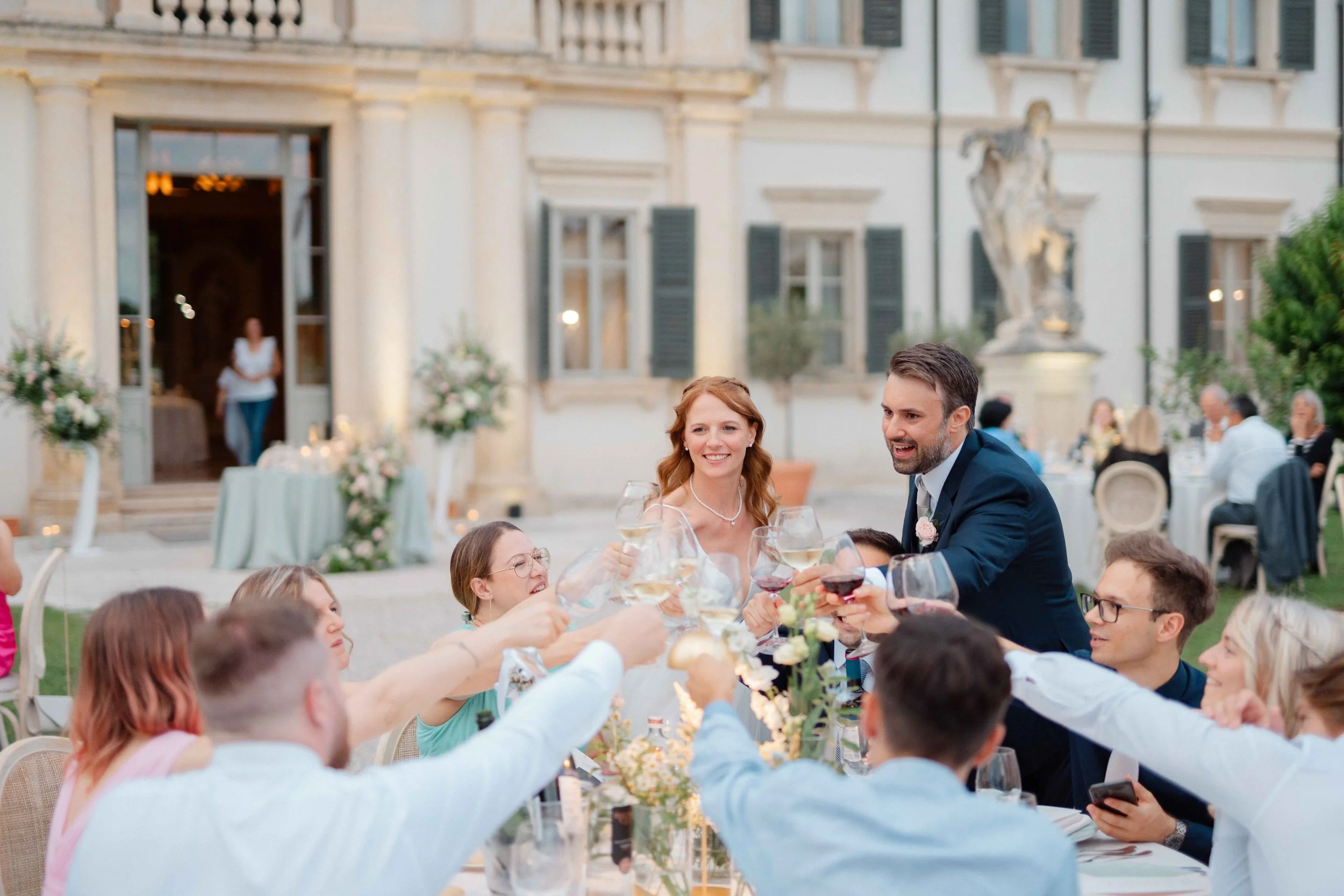 People raising glasses in celebration at an outdoor wedding or banquet dinner, with a large historic building in the background.