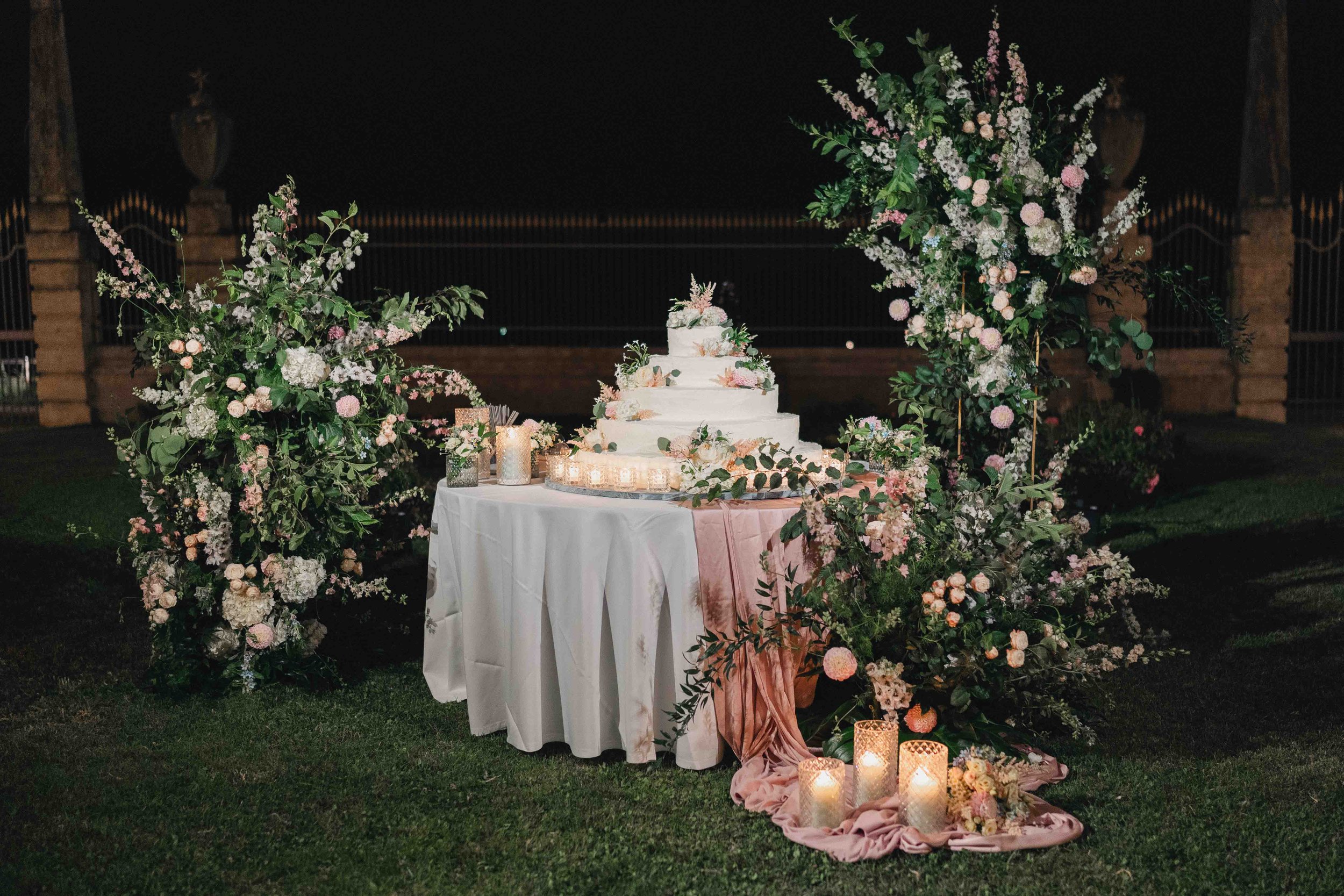 A wedding cake on a table decorated with pink and white flowers and surrounded by large floral arrangements, with candles on the ground, set outdoors at night.