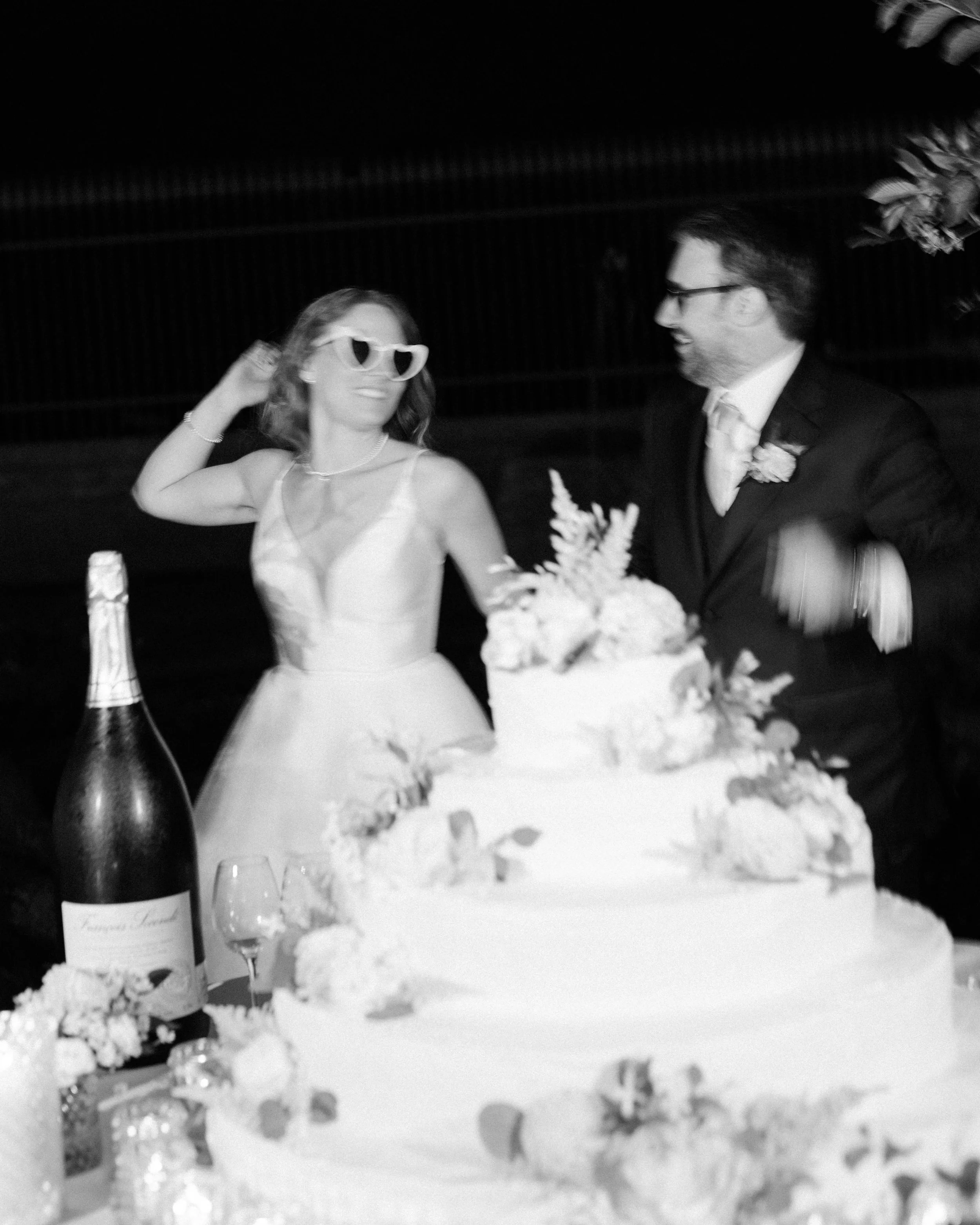 A black and white photo of a bride with sunglasses and a groom smiling at each other at a wedding reception. There is a wedding cake, a bottle of champagne, and floral decorations on the table.
