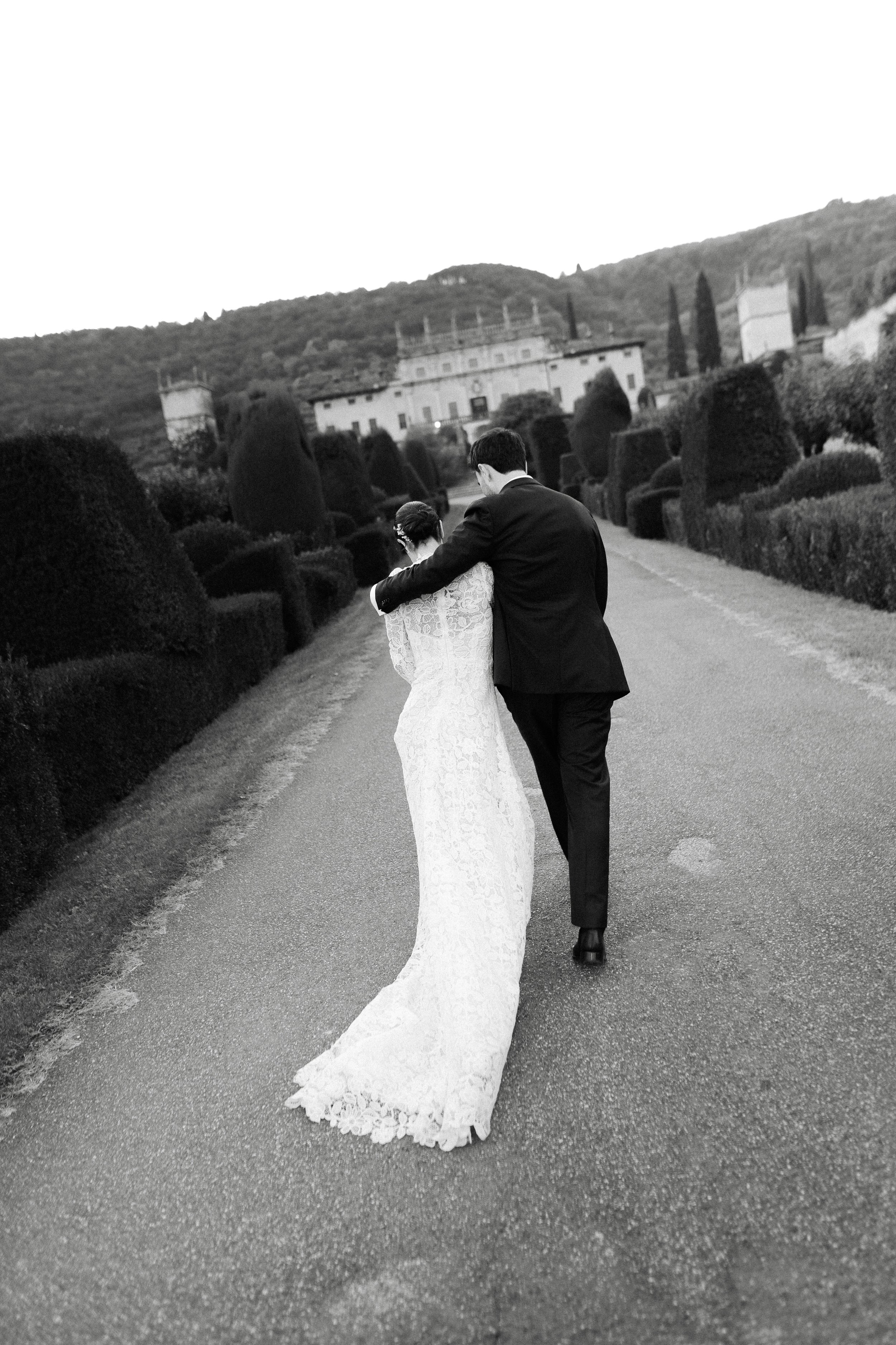 A bride and groom walking down a garden path with neatly trimmed bushes, with a castle-like building and hills in the background. The scene is in black and white.