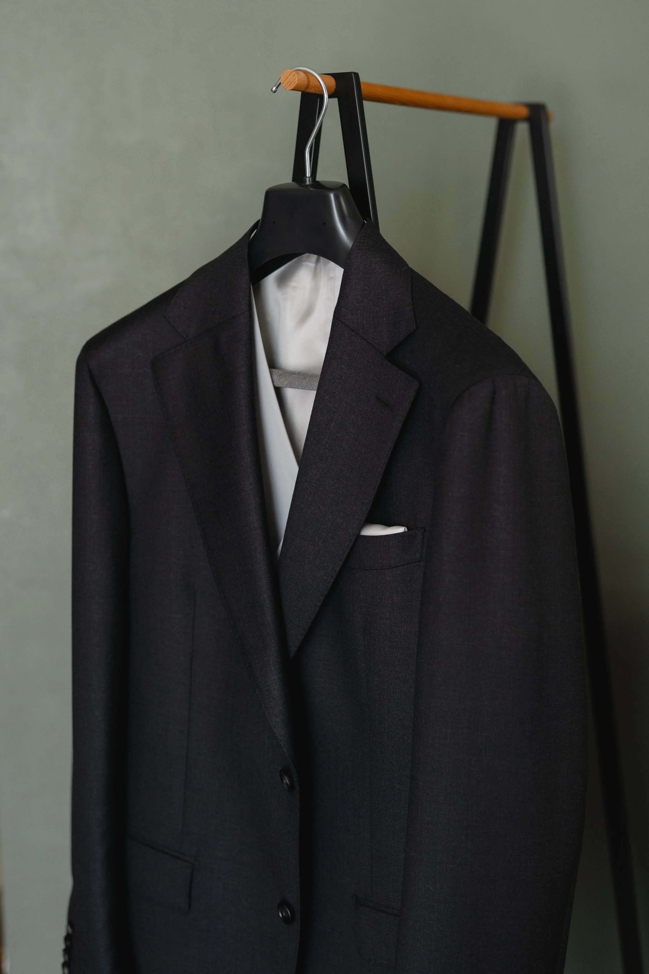 Black suit jacket hanging on a wooden and black metal clothing rack against a plain wall.