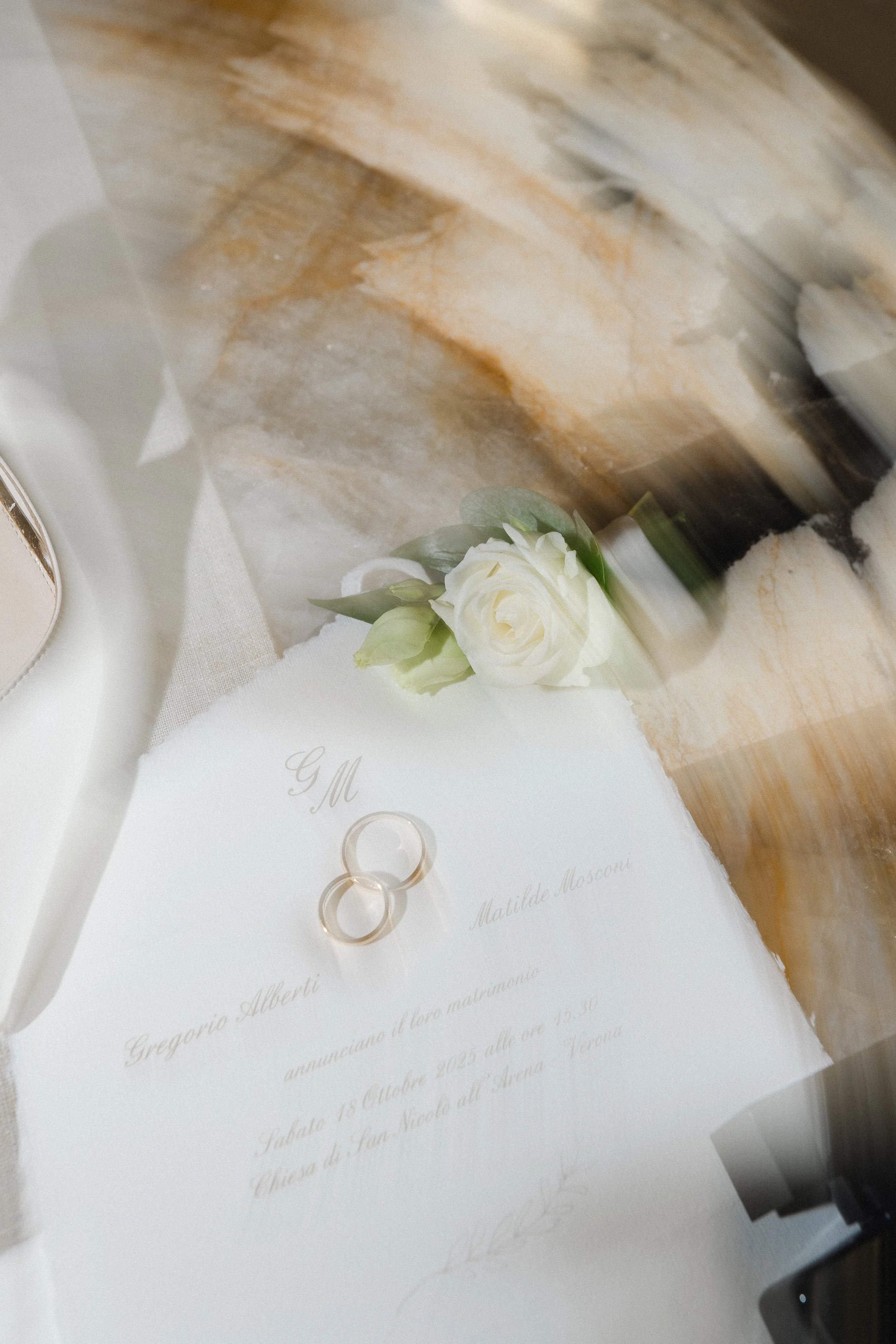 A wedding invitation card with gold rings and a white flower on it, partially visible, on a wooden surface.