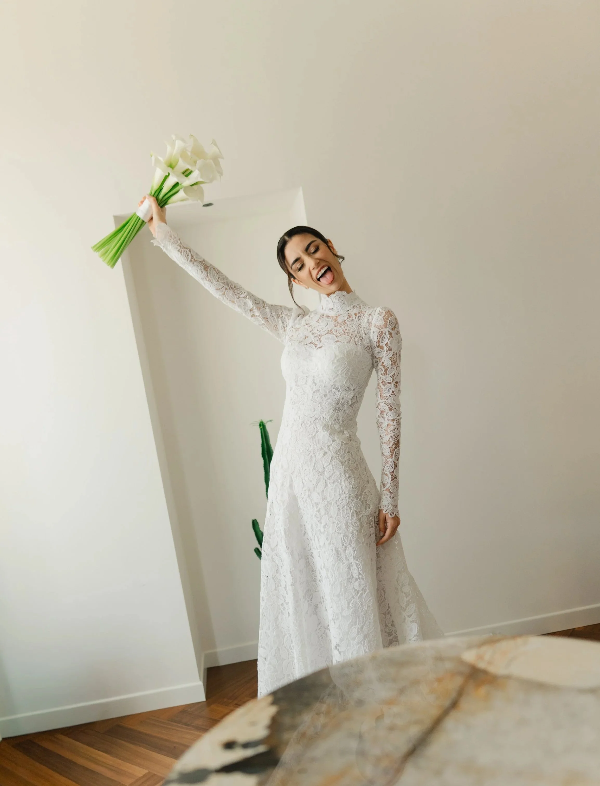 A woman in a long white lace dress holding a bouquet of white calla lilies, smiling and sticking out her tongue