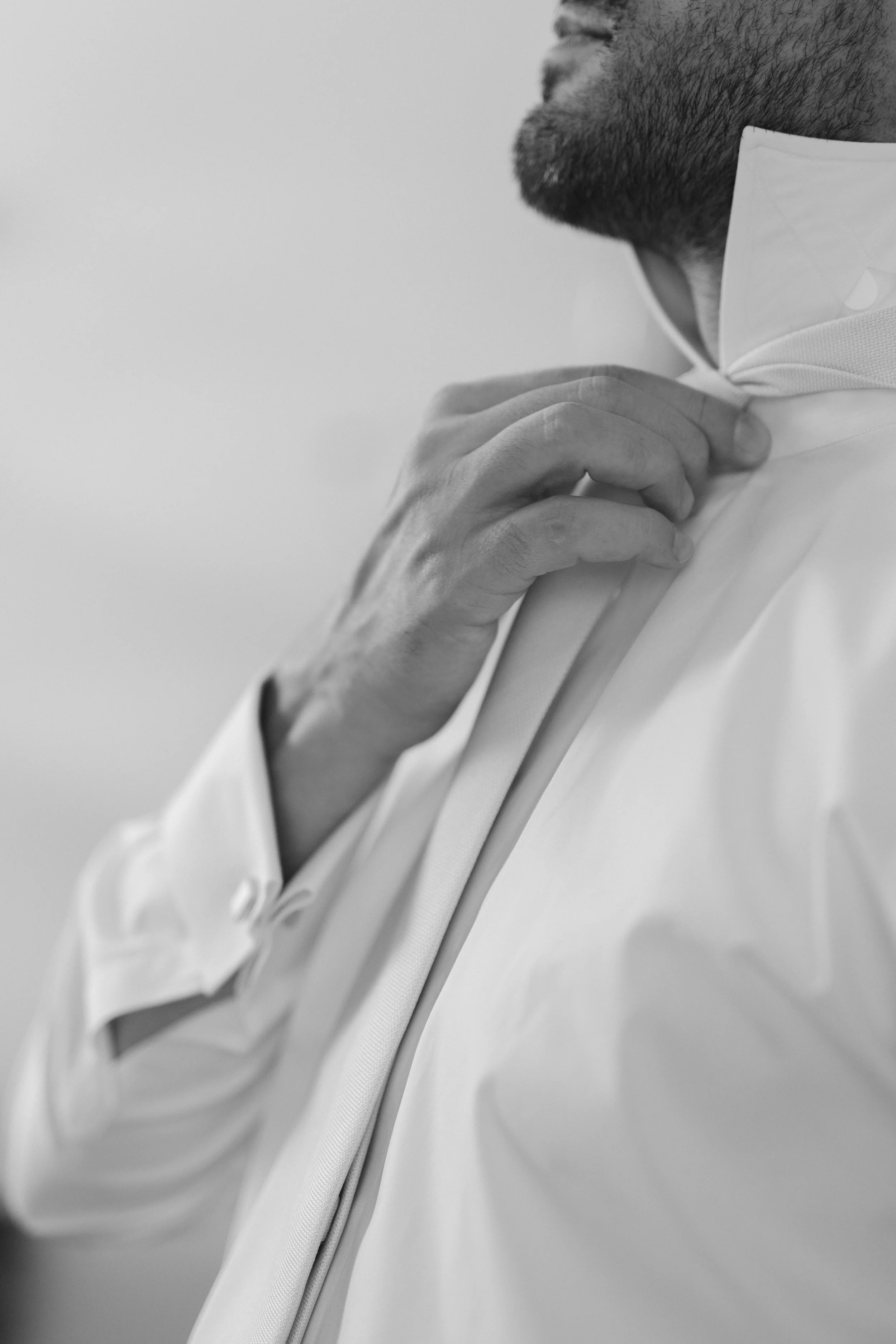 Close-up of a man adjusting a white shirt collar. He has dark, short hair and a beard, and is wearing a medical face mask. The background is plain and blurred.