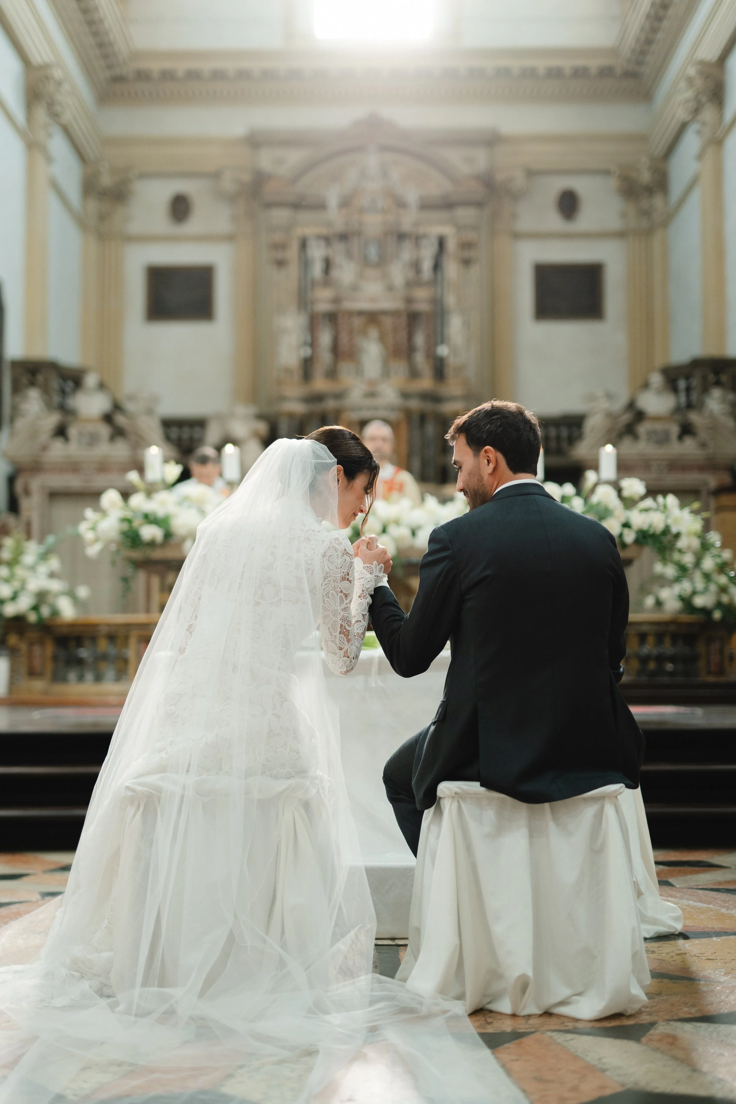 A bride and groom kneeling during a wedding ceremony in a church, holding hands, with an altar decorated with flowers in the background.