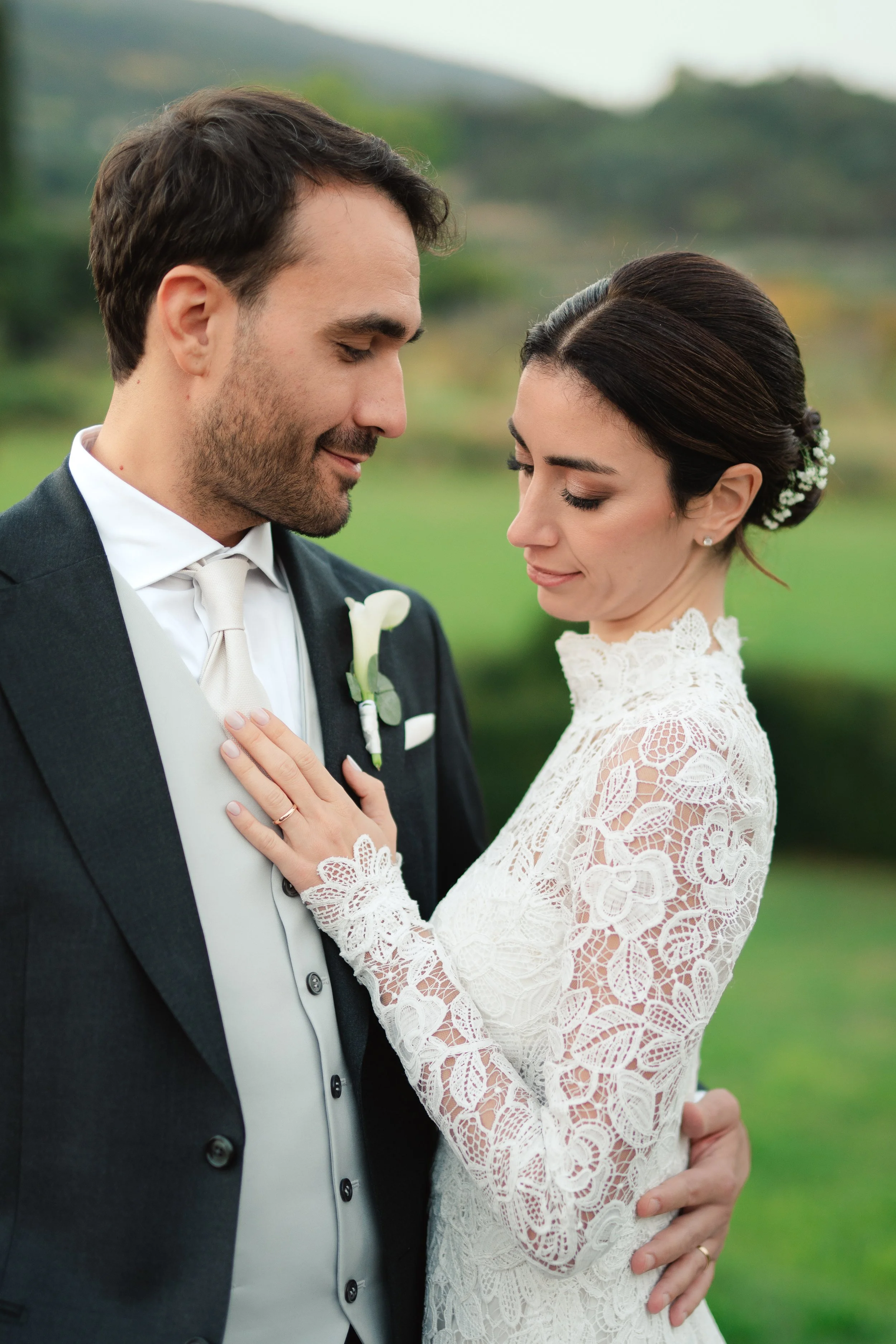 A bride and groom stand close to each other outdoors, with the bride holding the groom's chest and the groom touching the bride's waist, both with gentle smiles. The groom wears a dark tuxedo with a white shirt and tie, and the bride wears a white lace wedding dress with floral hair accessories.