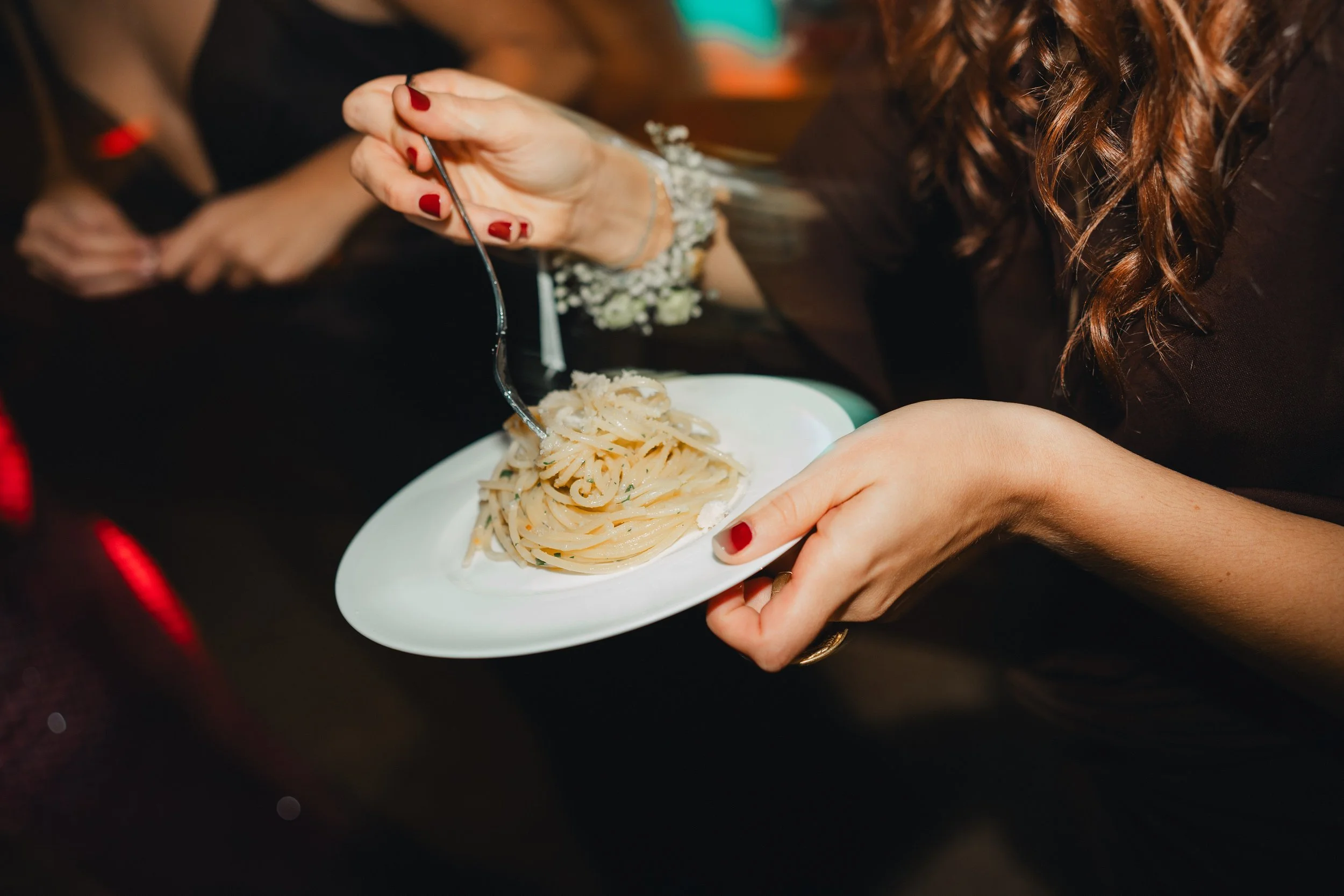 A woman with red nail polish and a floral bracelet is holding a spoon and a white plate with spaghetti pasta, in a setting with blurry background.