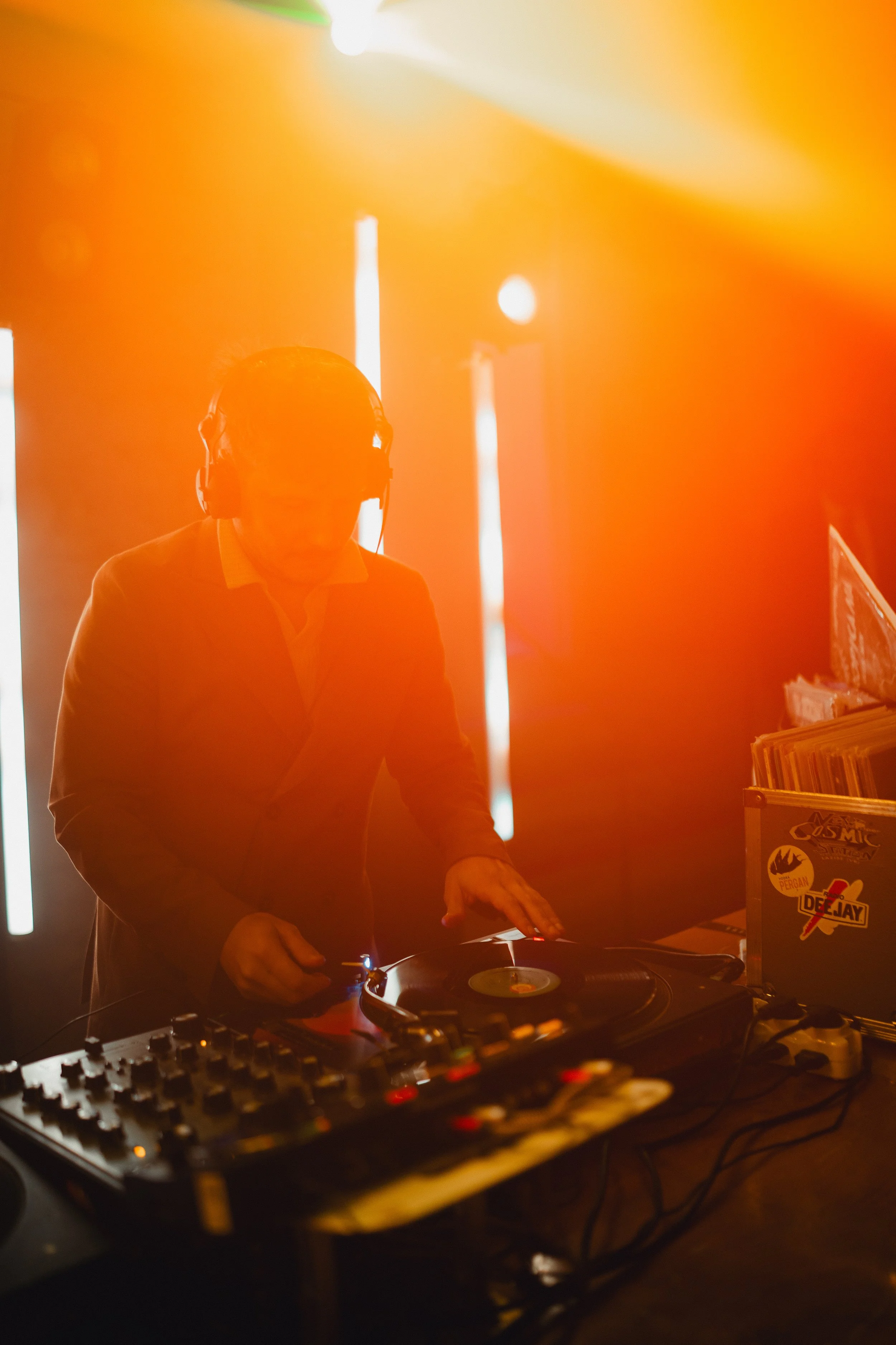 A DJ wearing headphones and dark clothing is playing music on a turntable and mixer in a dimly lit room with orange lighting.