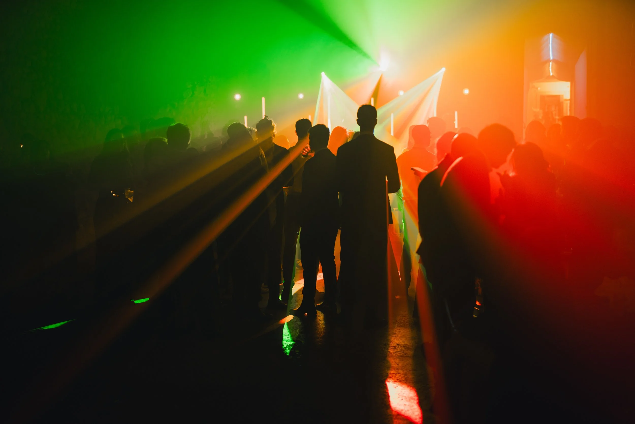 People in a dark room with colorful laser lights, standing and watching a light show or performance.