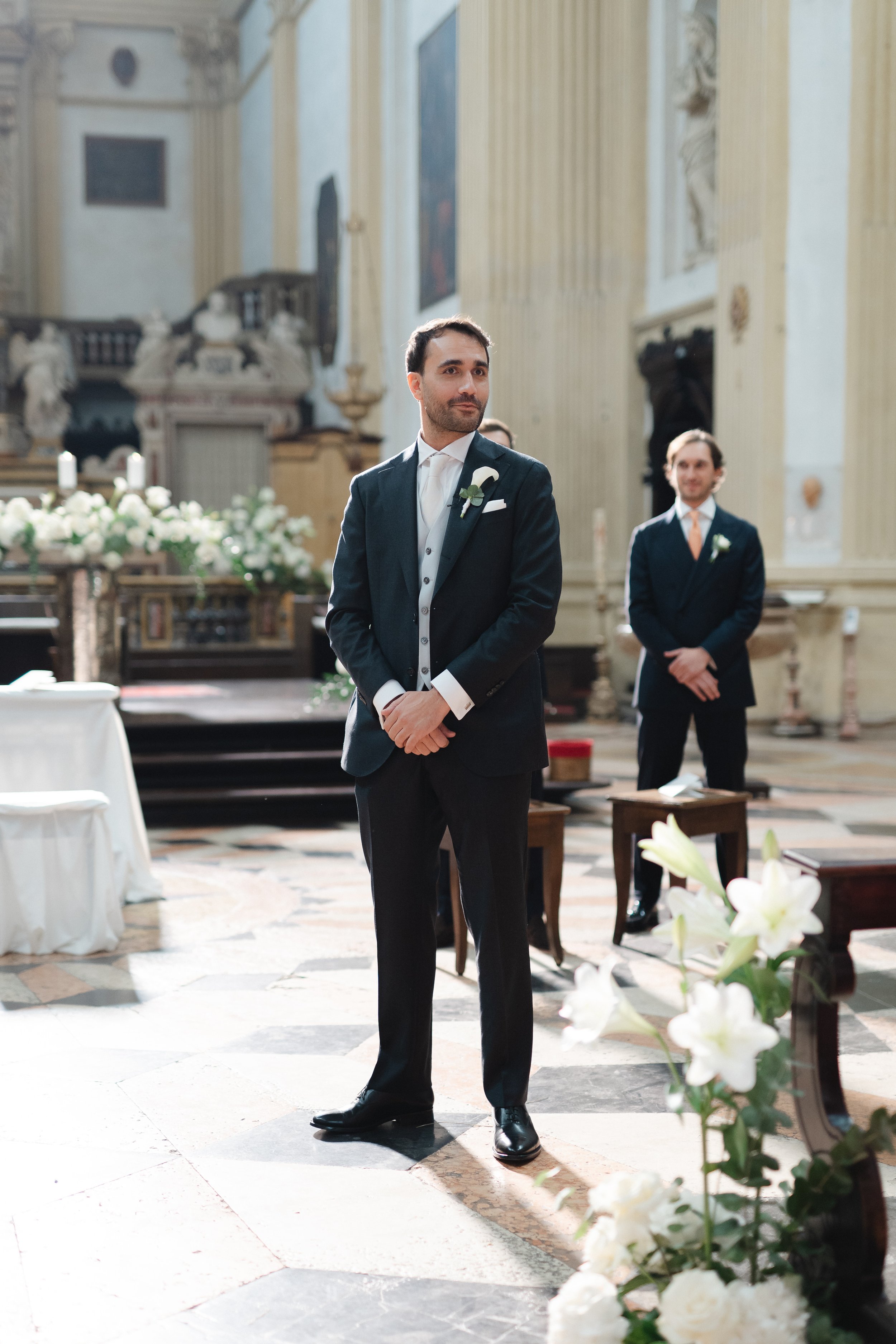 A groom in a dark suit and white tie stands inside a church, looking to his right. Behind him, another man in a dark suit with an orange tie stands with his hands clasped. The church has ornate decor, religious paintings, and white flower arrangements.