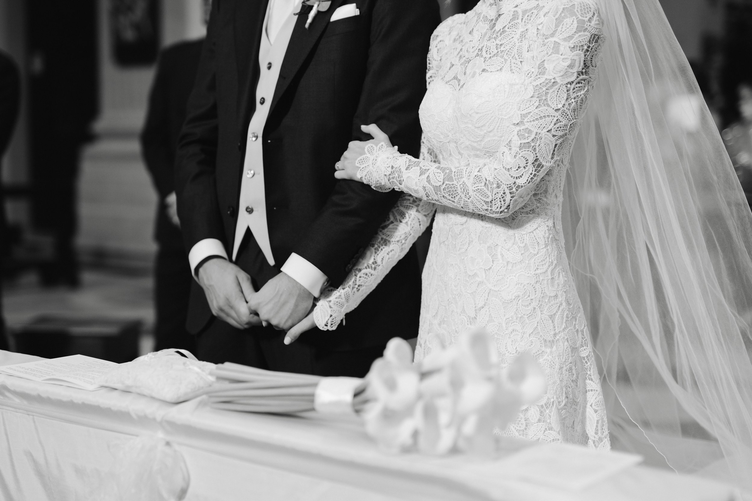 A bride and groom holding hands during a wedding ceremony, with the bride wearing a lace wedding dress and a veil, and the groom dressed in a black suit with a white shirt, standing at a table covered with a white cloth.