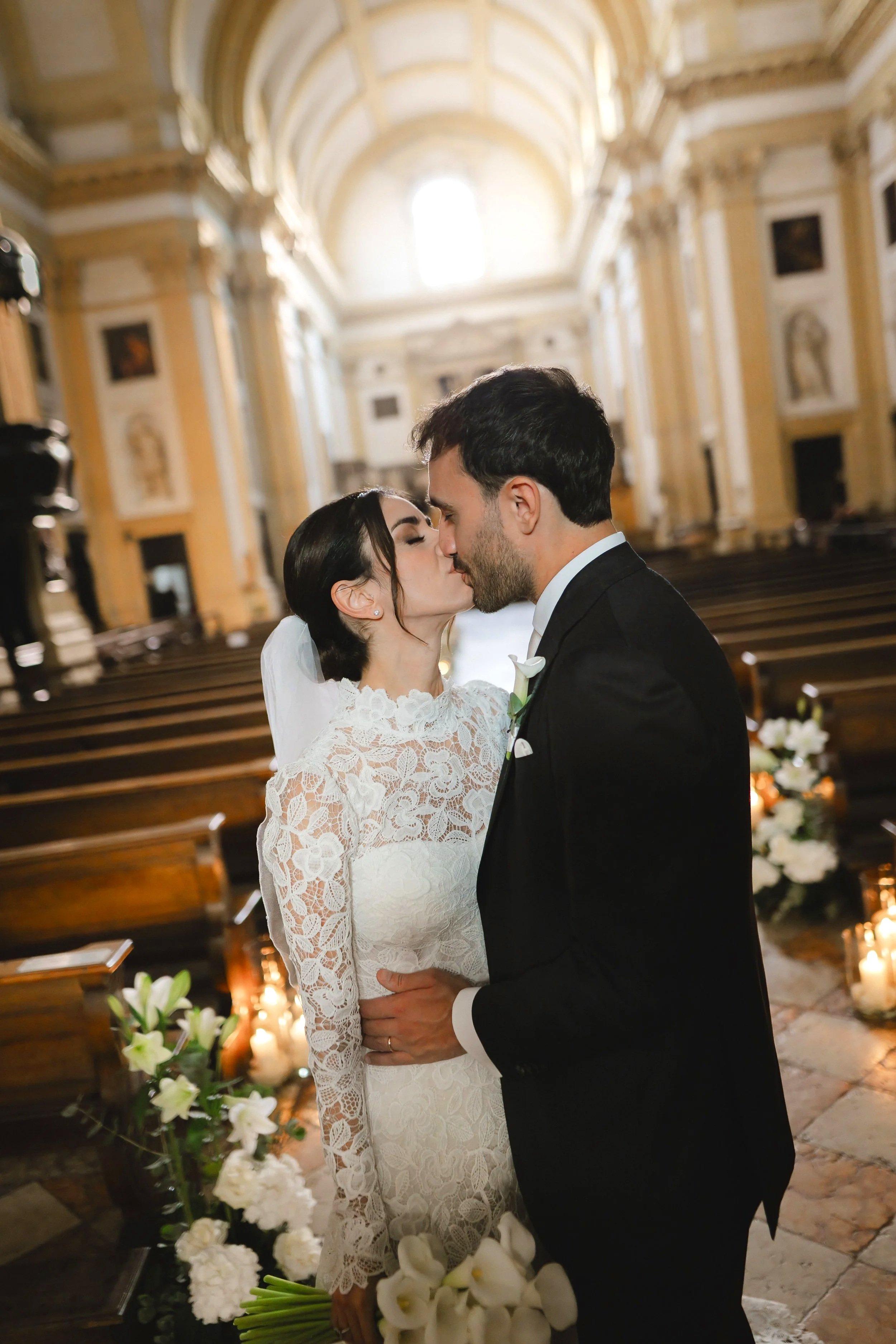 A bride and groom kiss inside a church. The bride is wearing a lace wedding dress and veil, holding a bouquet of white flowers. The groom is in a black tuxedo. The church has ornate gold and white decor with floral arrangements and candles.