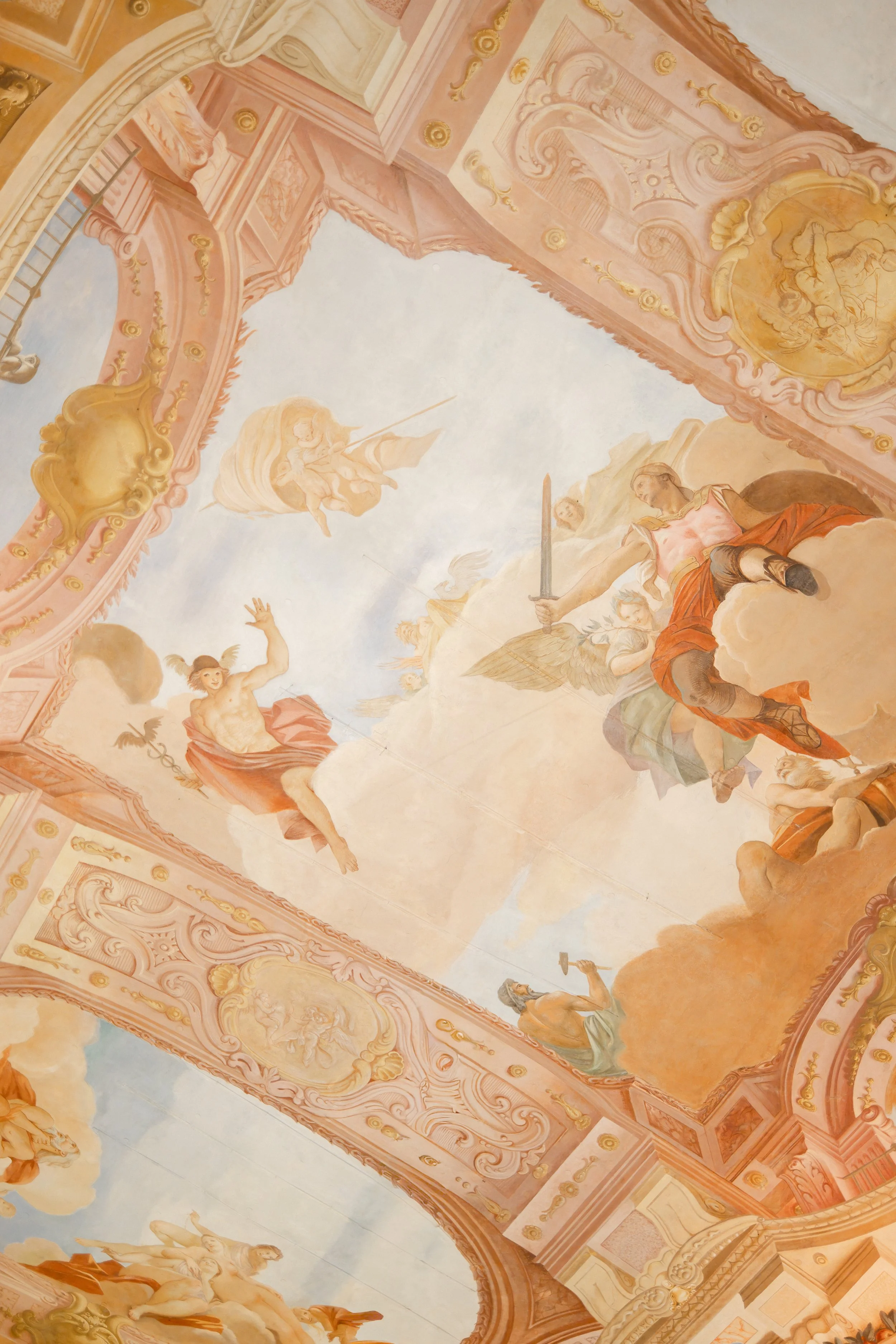 Baroque style ceiling painting with cherubs, angels, and mythological figures amid ornate gold and pastel pink decorative elements.