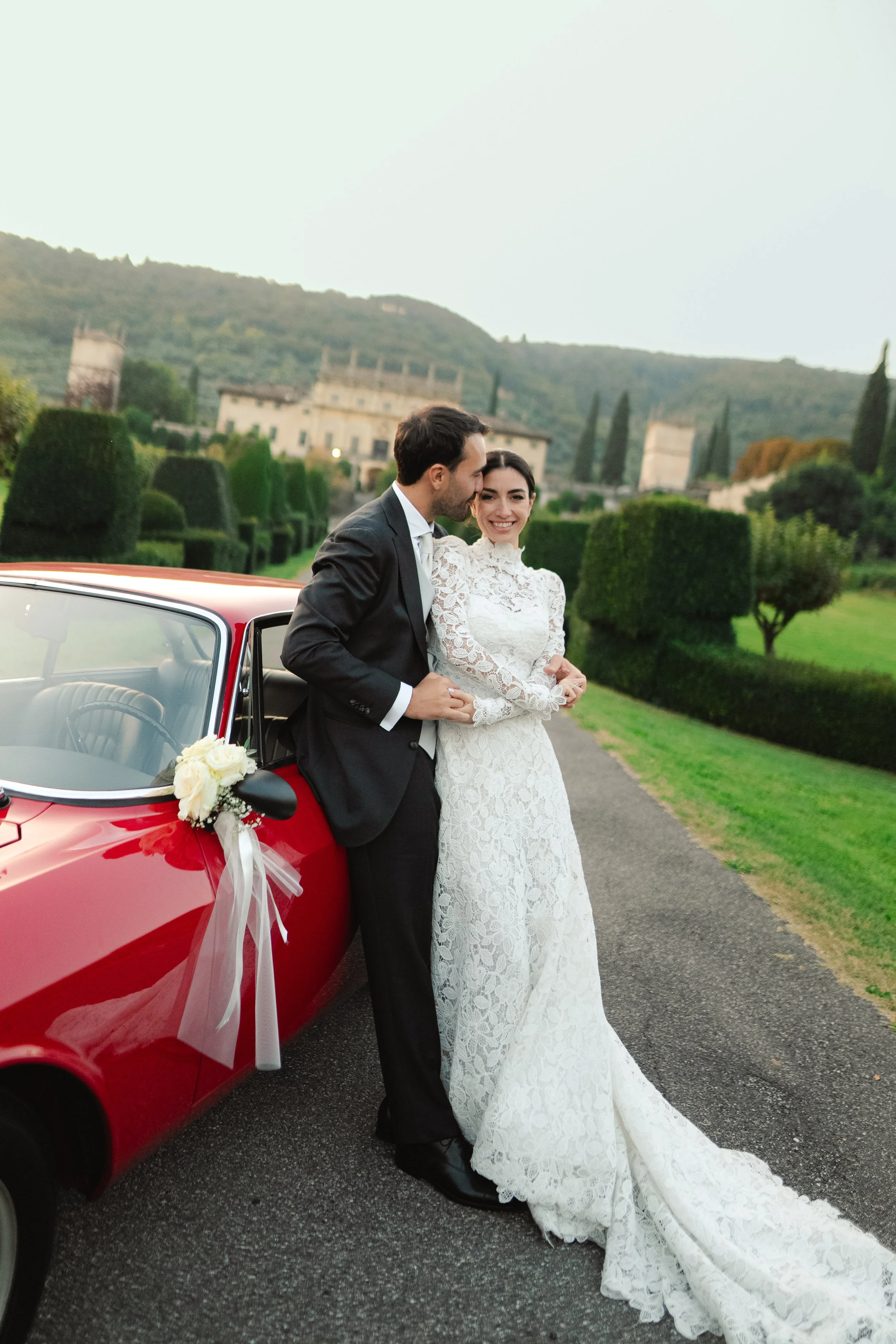 A newlywed couple stands beside a red vintage car decorated with white ribbons and flowers, in an outdoor garden setting with a historic building and hills in the background.