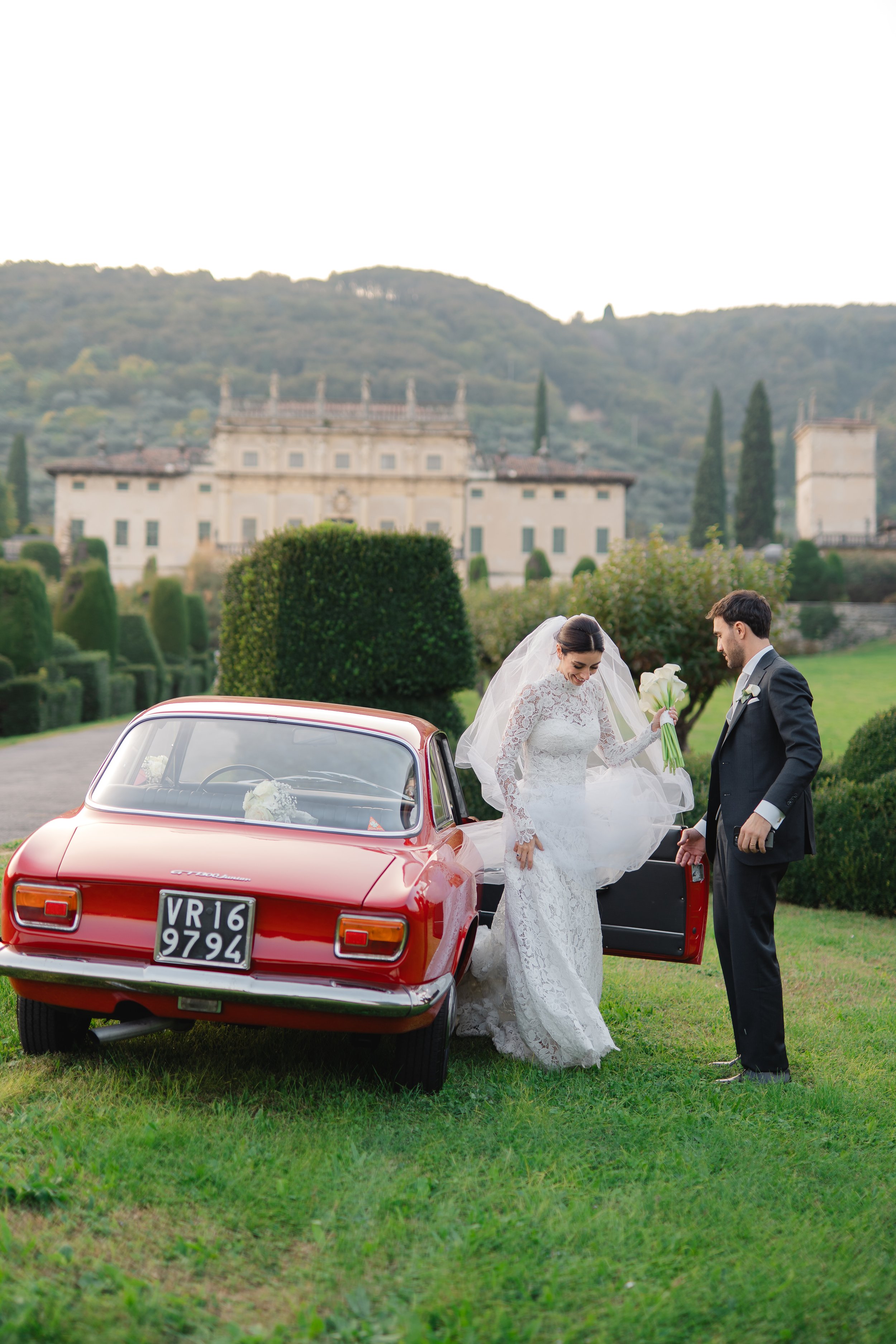 Bride and groom standing outdoors near a red vintage car, in front of Villa Arvedi.