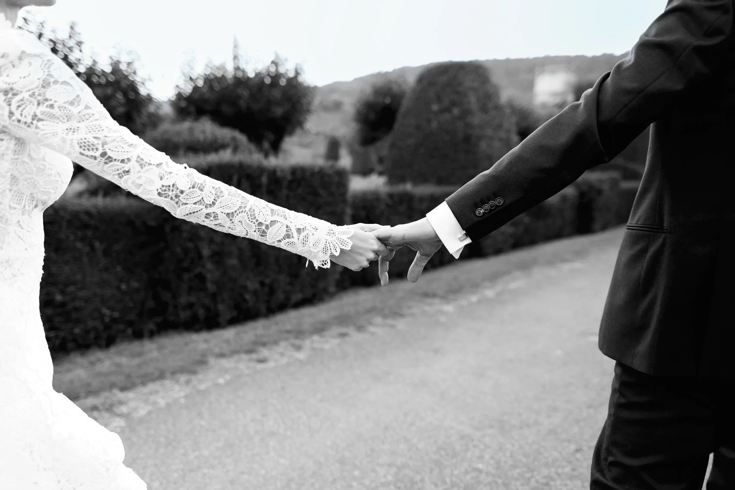 A black and white photo of a bride and groom holding hands outdoors. The bride is wearing a lacy long-sleeved dress, and the groom is in a dark suit. Their hands are clasped, symbolizing a wedding or romantic moment.