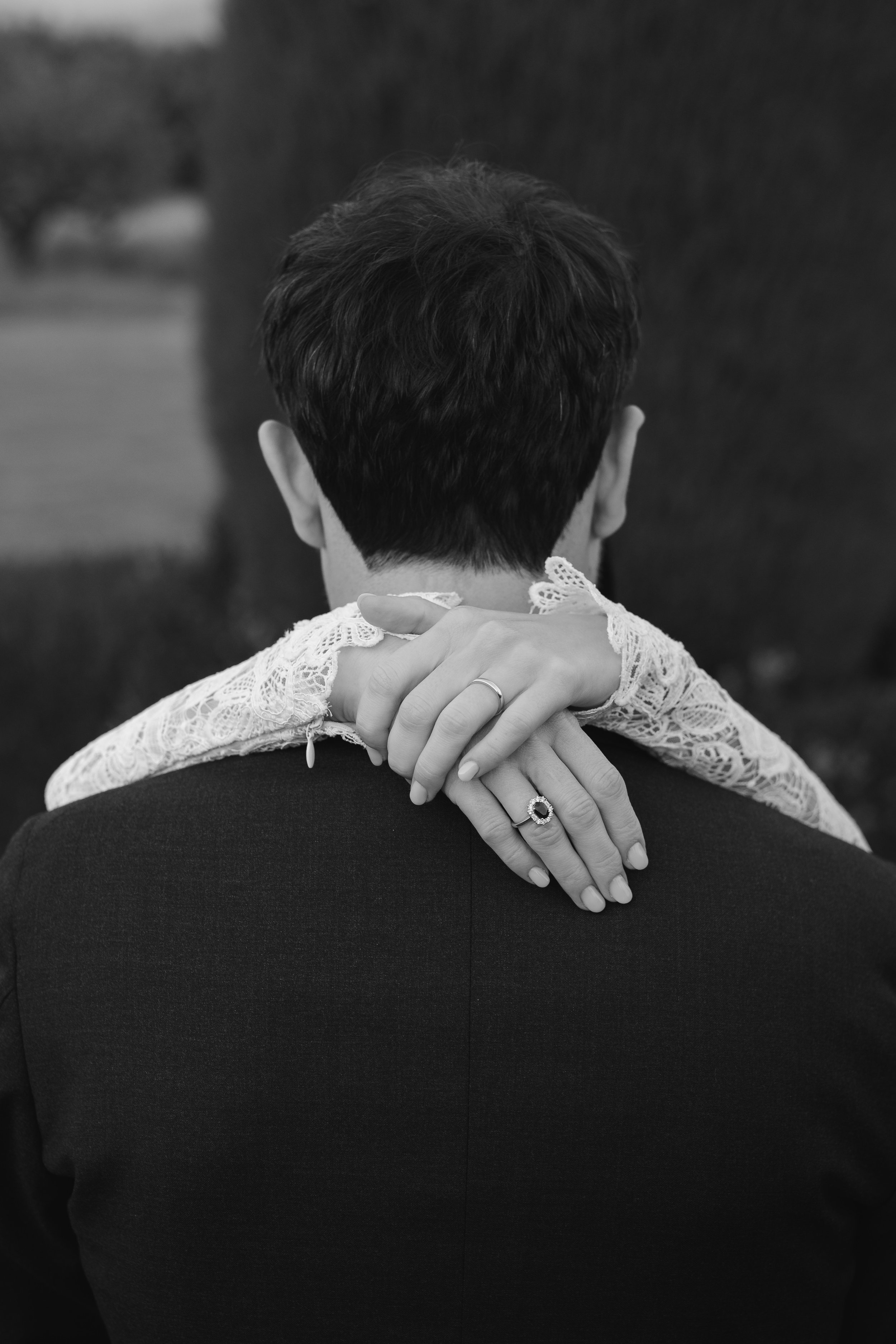 Black and white photograph of a woman and a man, with the woman resting her arms on the man's shoulders from behind, showing her hands with rings, and the back of the man's head and shoulders.