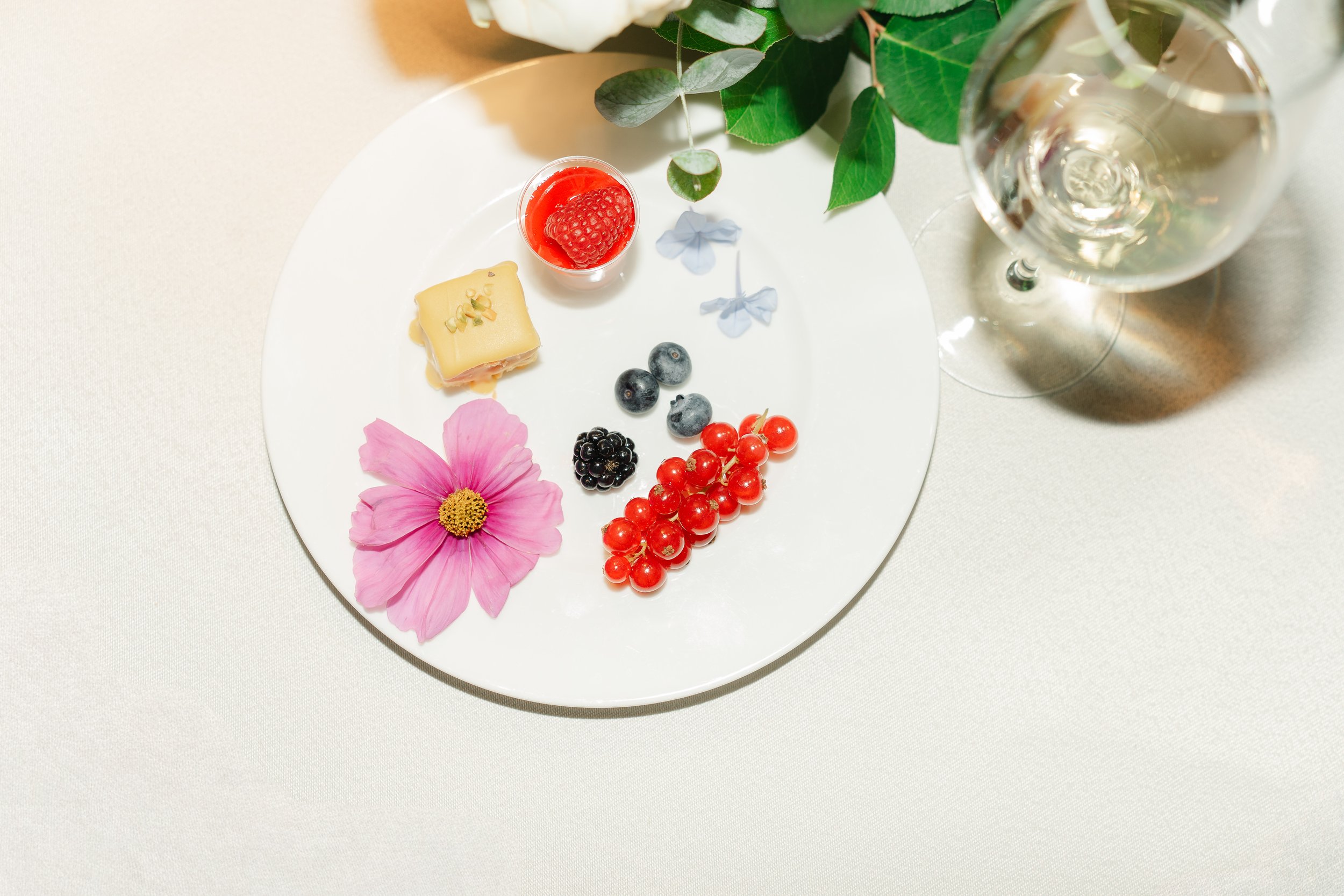 A white plate with a pink flower, a small square of cheese topped with chopped nuts, a cluster of red currants, blueberries, blackberries, a small cup of red jelly with a raspberry, and two white butterflies. A wine glass with water and green leaves 
