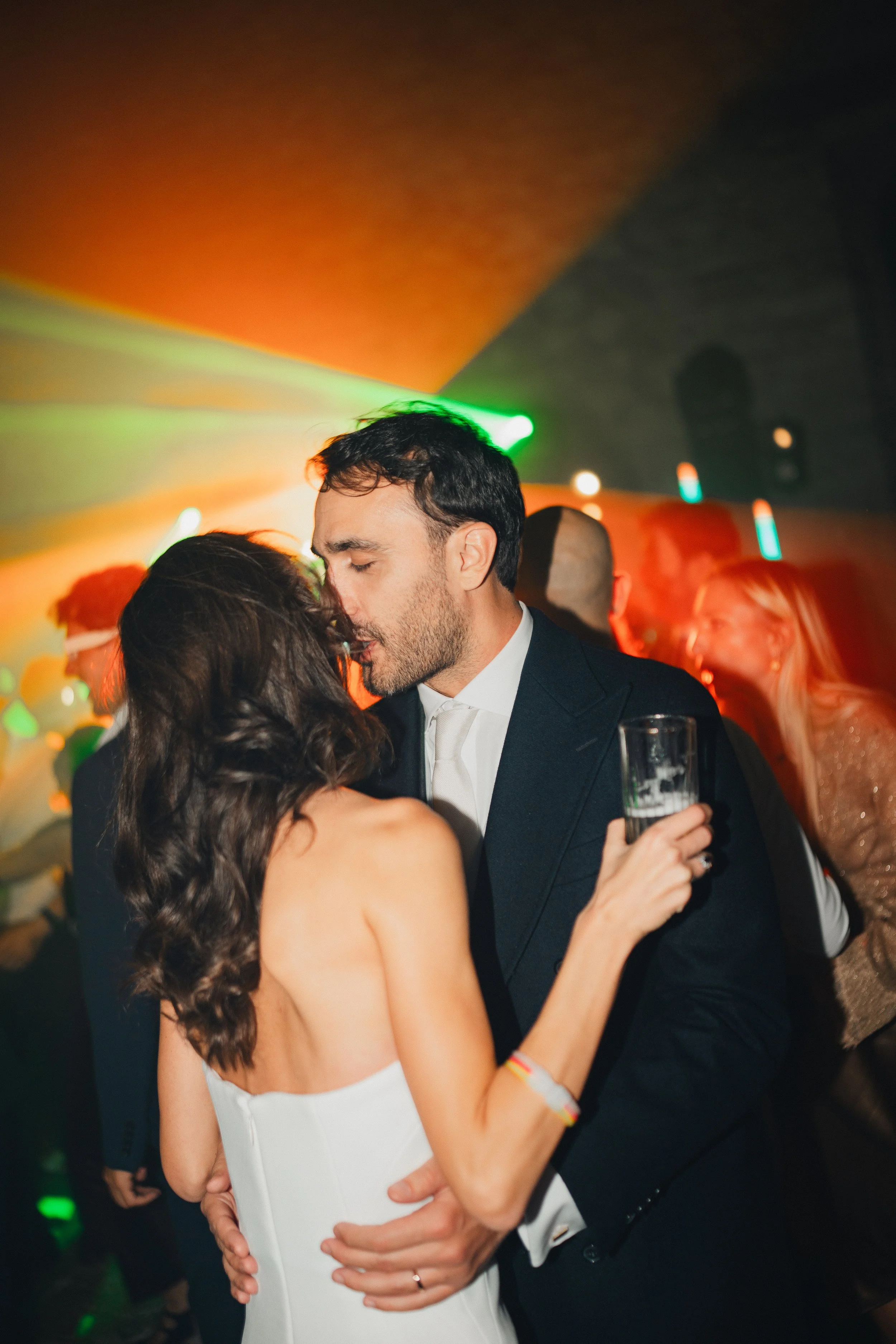 A man in a suit and a woman in a white dress are dancing closely at a party or wedding reception. The woman has long, dark hair and the man has short, dark hair and a beard. The woman is holding a glass of drink, and they are surrounded by other guests in a dimly lit venue with colorful lights.