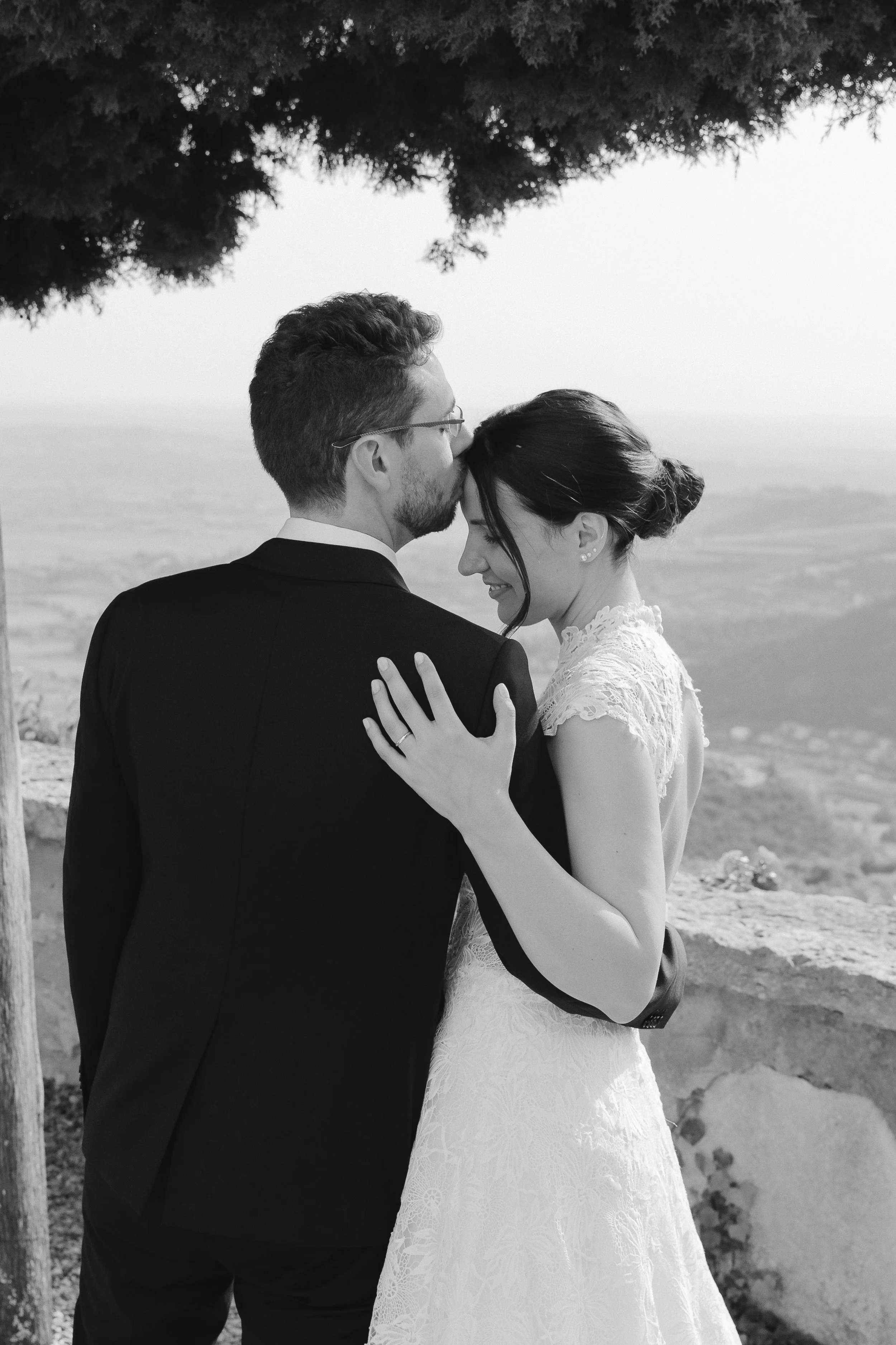 A black-and-white photograph of a bride and groom embracing outdoors, with the groom kissing the bride's forehead, both smiling and holding each other gently. The background features landscape and a large tree overhead.