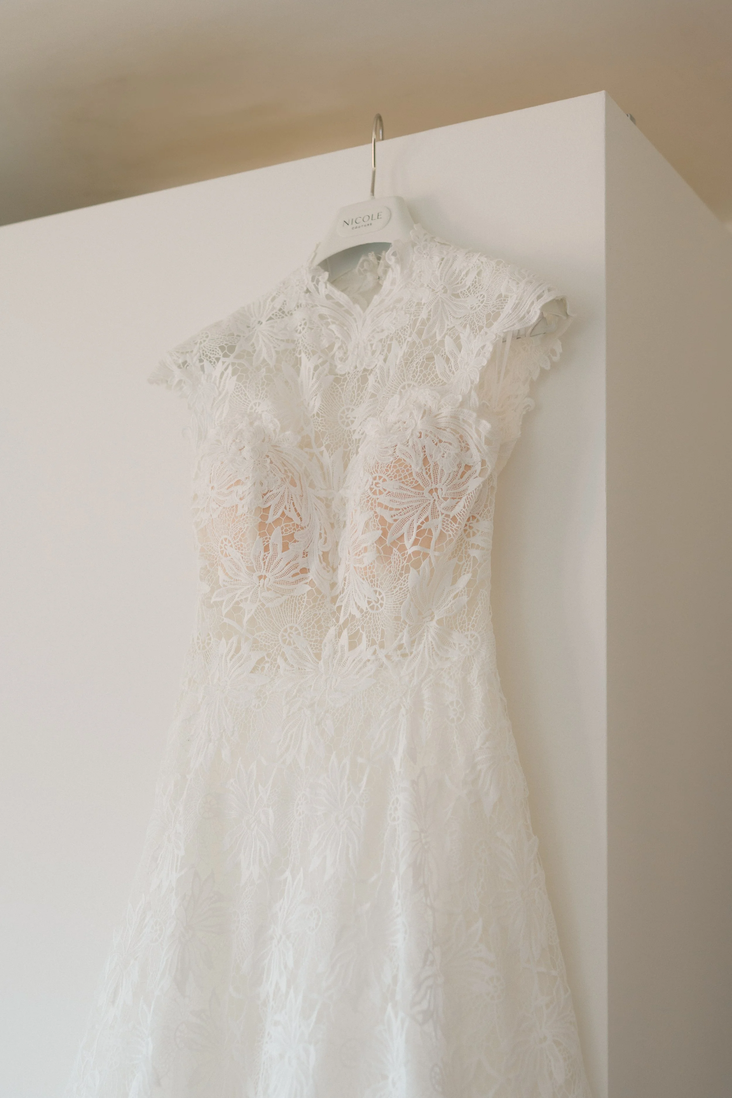 A white lace wedding dress hanging on a white hanger with the name Nicole, on a white wall.