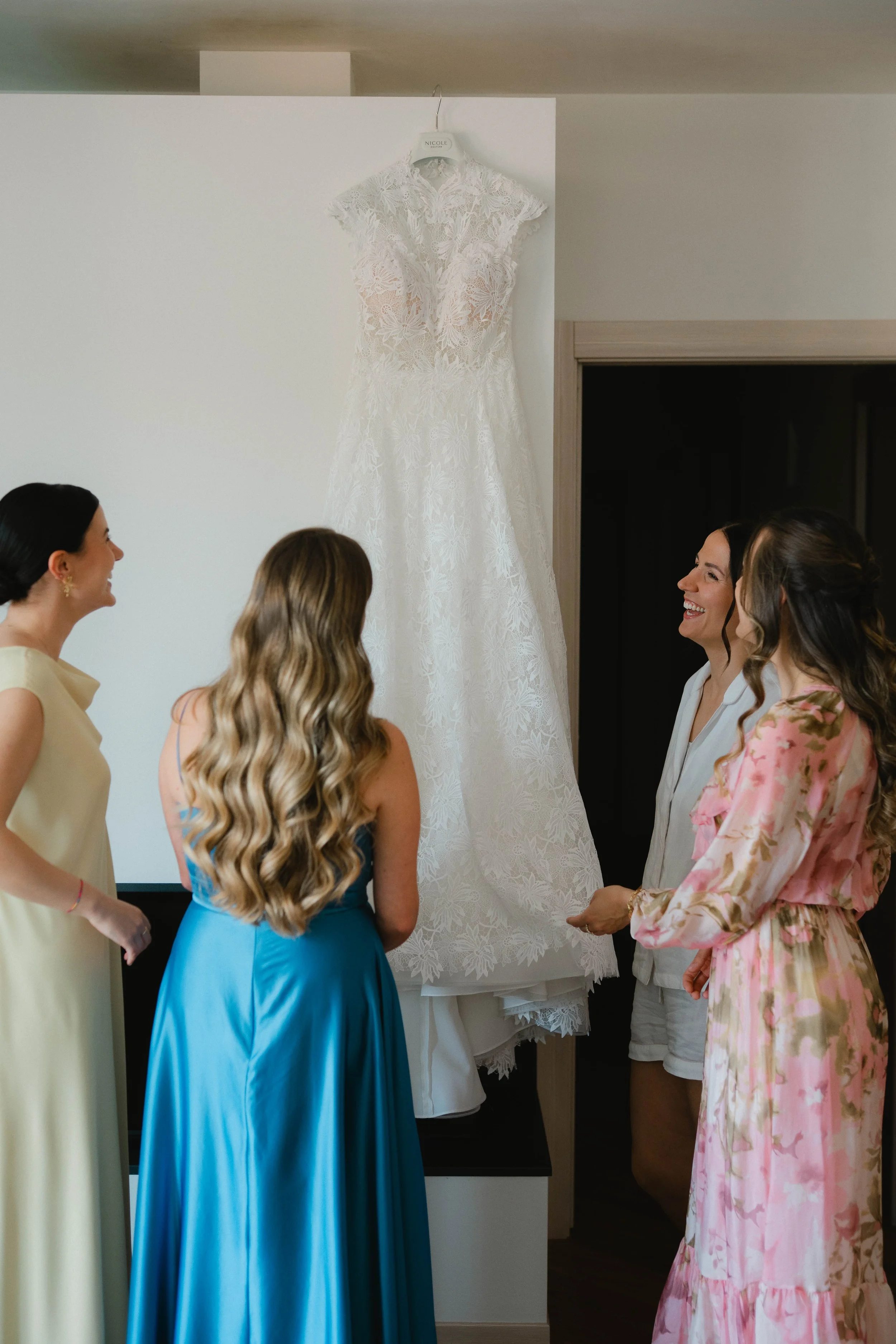 A group of women in colorful dresses are gathered and smiling, nearby a white lace wedding dress hanging on a wall.