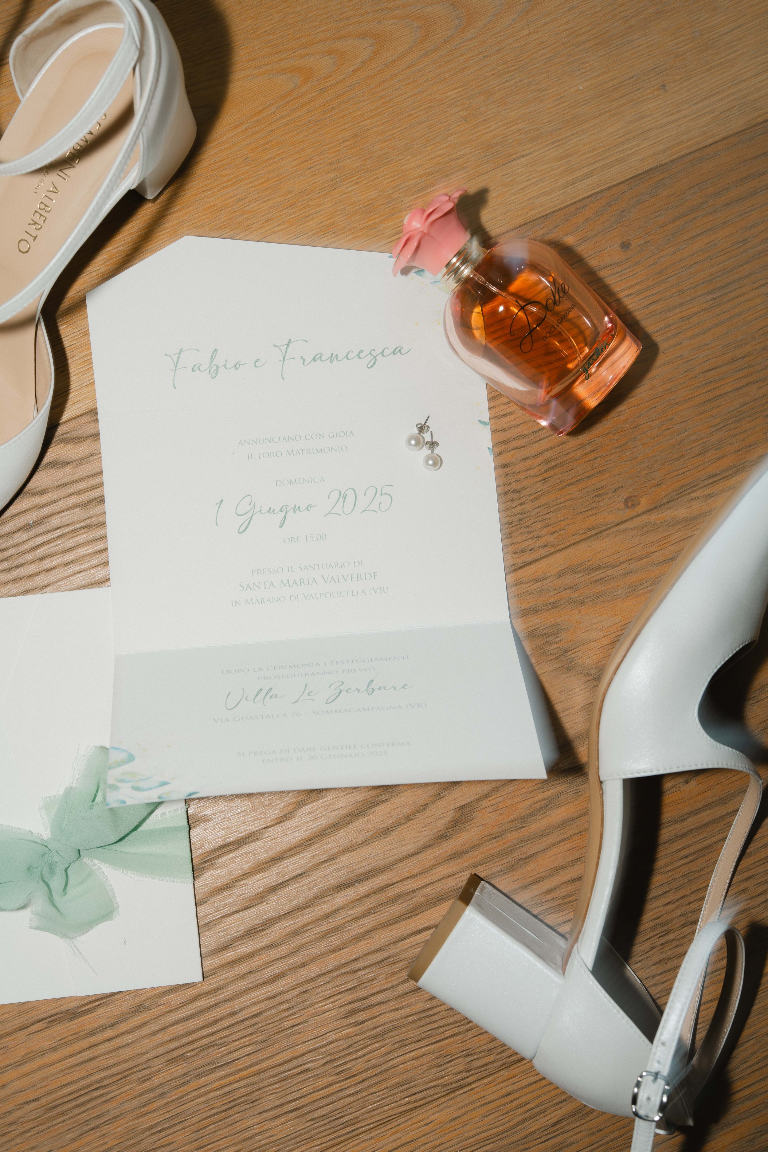 Wedding invitation, a perfume bottle, and a pair of white shoes on a wooden table, with a white chair nearby.