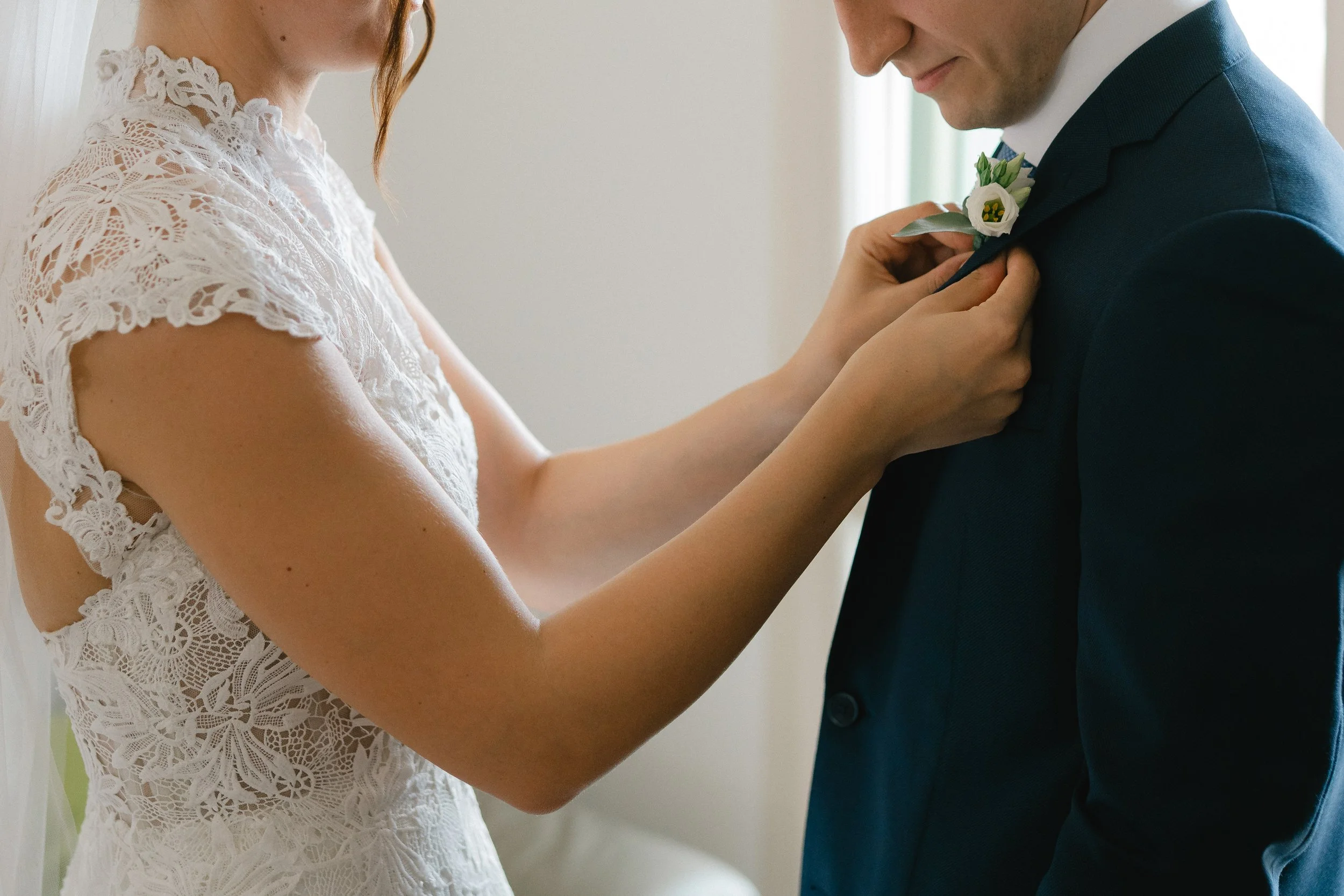A bride and groom standing close, with the bride adjusting the groom's boutonniere on his suit jacket. The bride wears a white lace dress, and the groom is dressed in a dark suit.