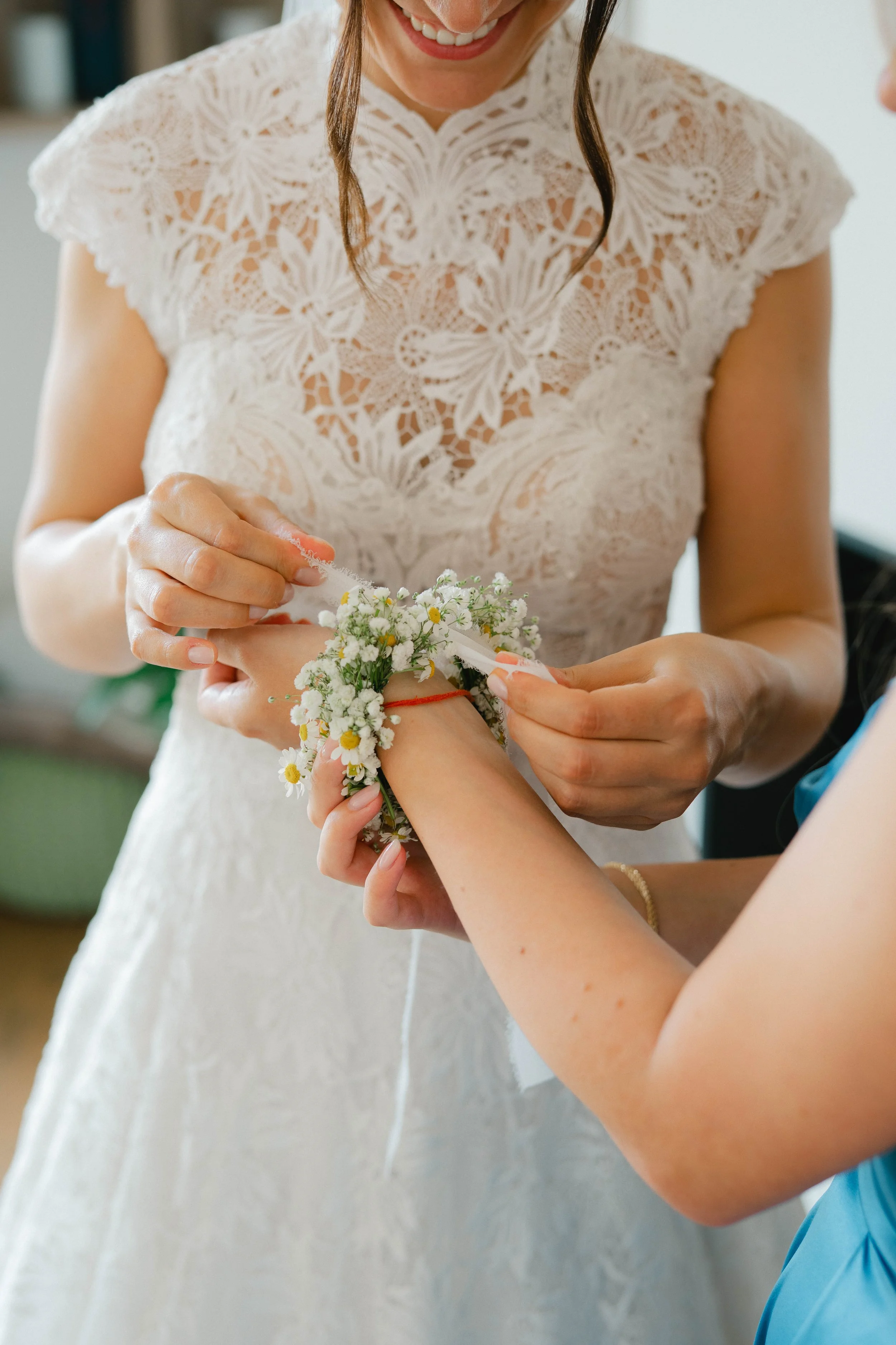 A woman in a white lace dress is smiling as she helps another woman put a flower bracelet on her wrist.