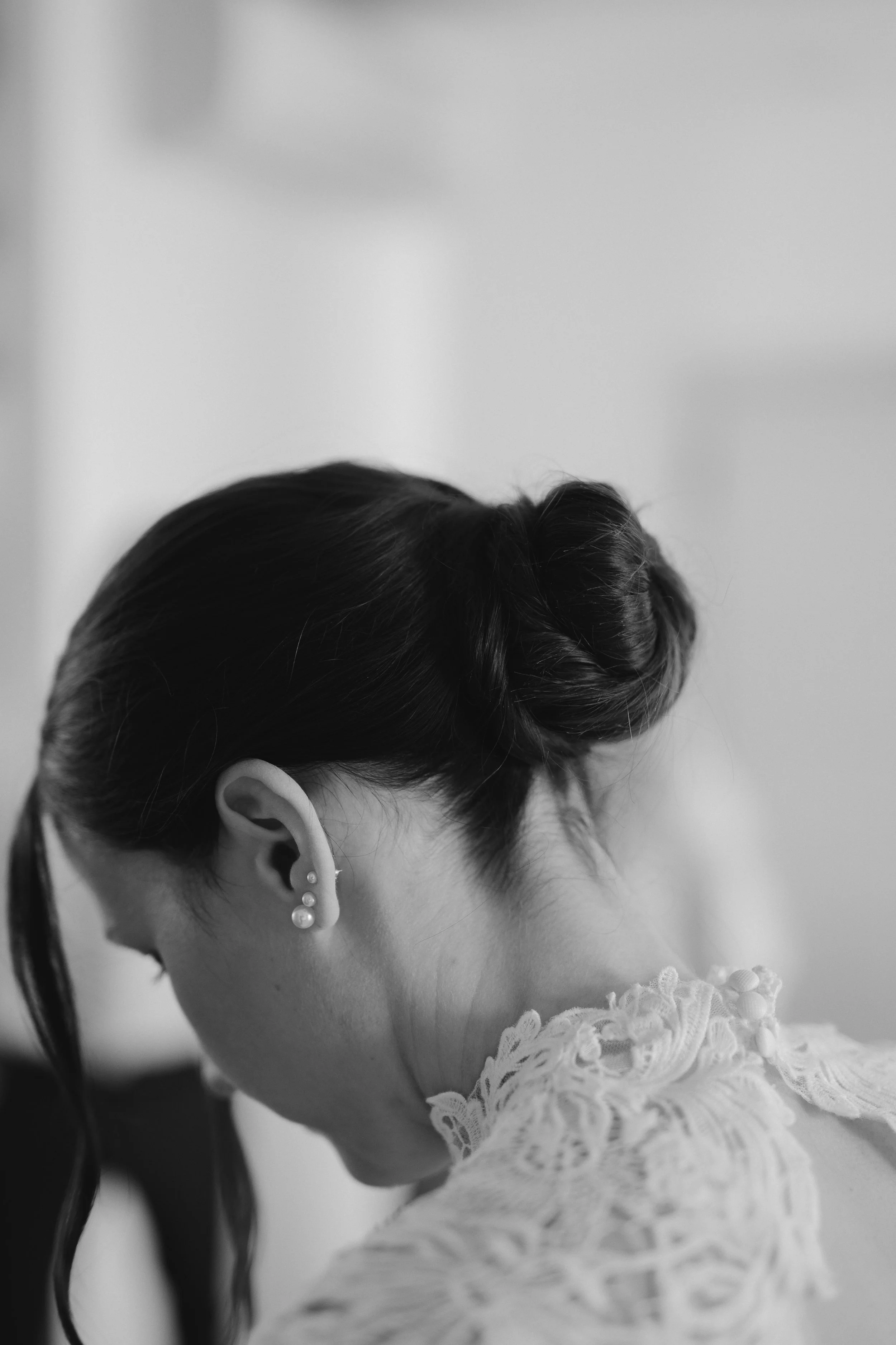 Close-up black and white photo of a young girl with dark hair styled in a neat updo, wearing pearl earrings and a lace dress, looking downward.