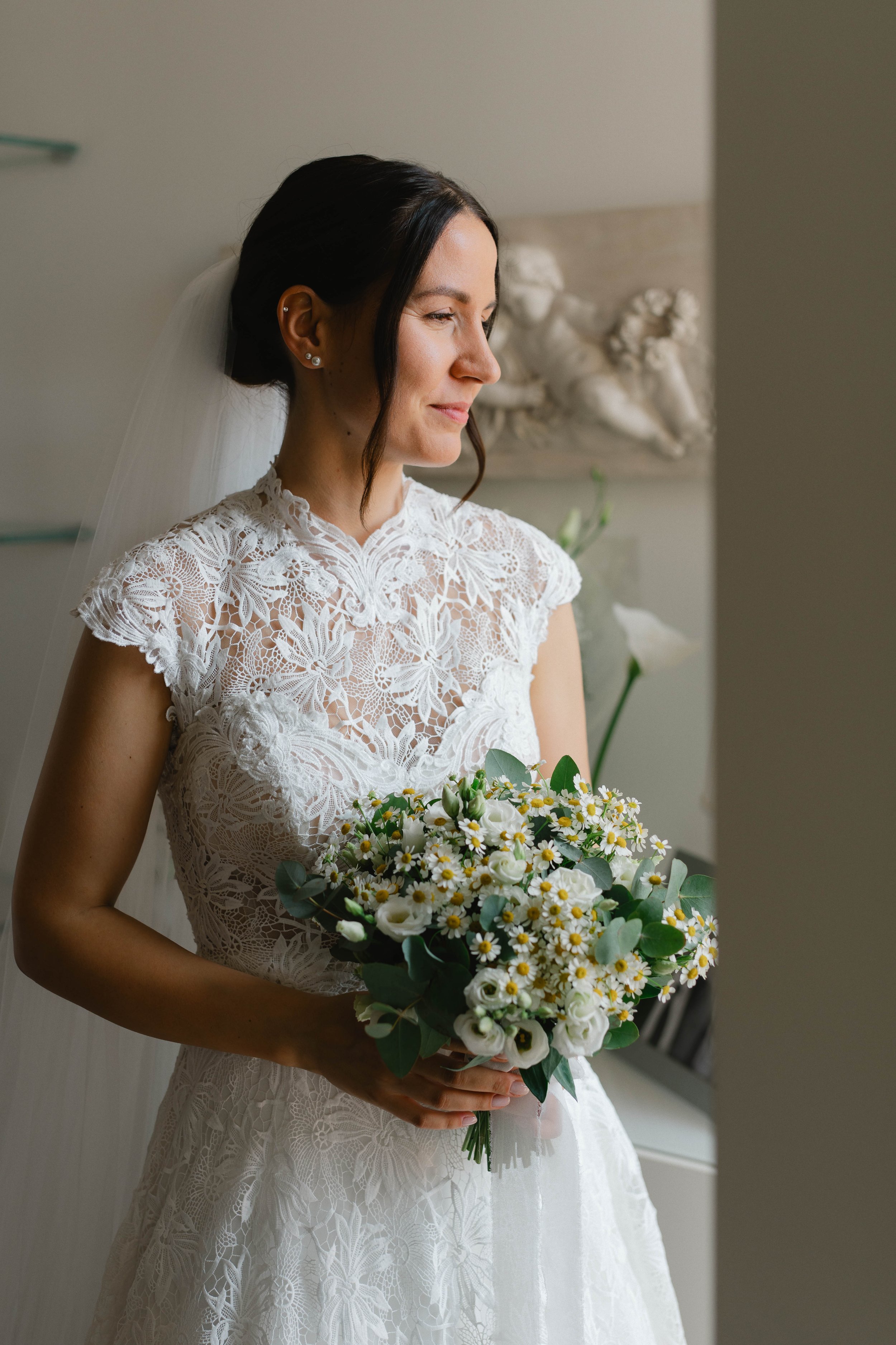 A bride in a lace wedding dress holding a bouquet of white flowers and greenery, standing indoors with a neutral background.