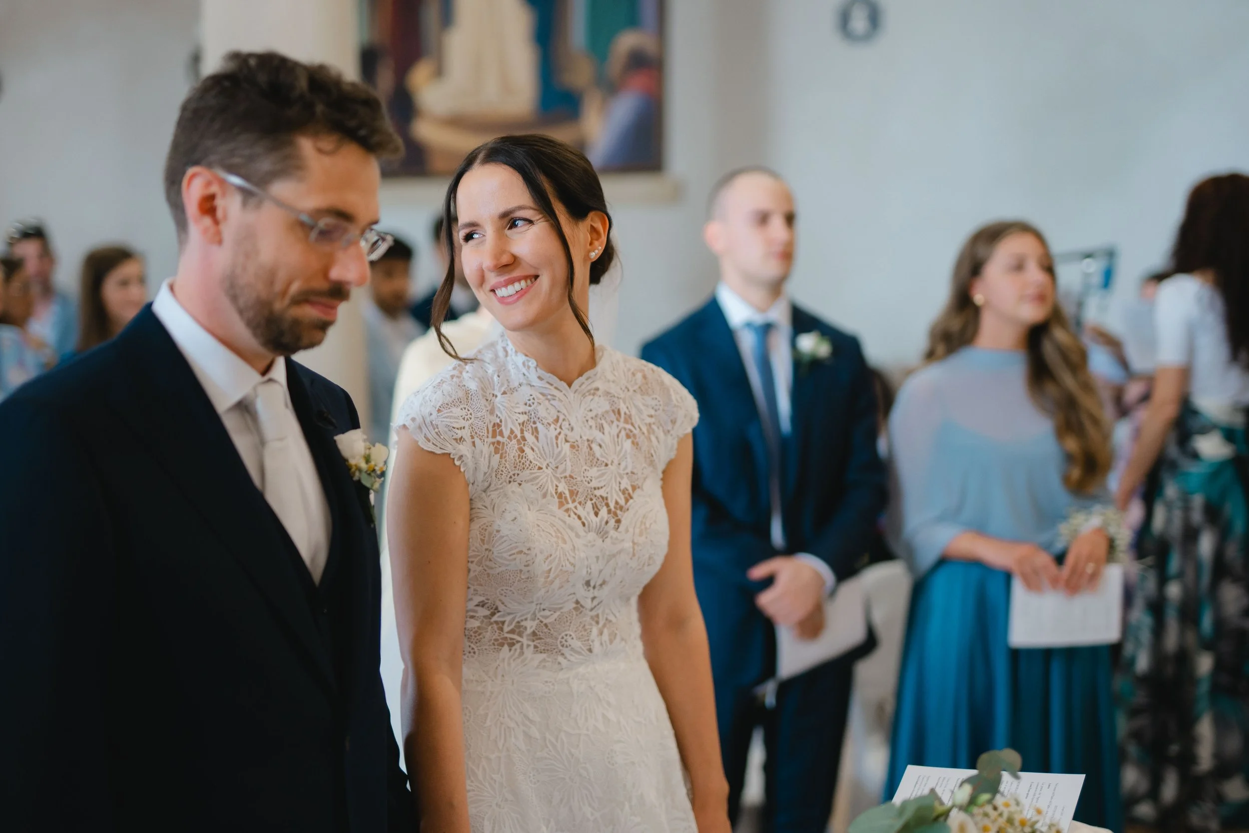 A bride and groom standing side by side at a wedding ceremony, surrounded by guests, in a bright indoor setting.