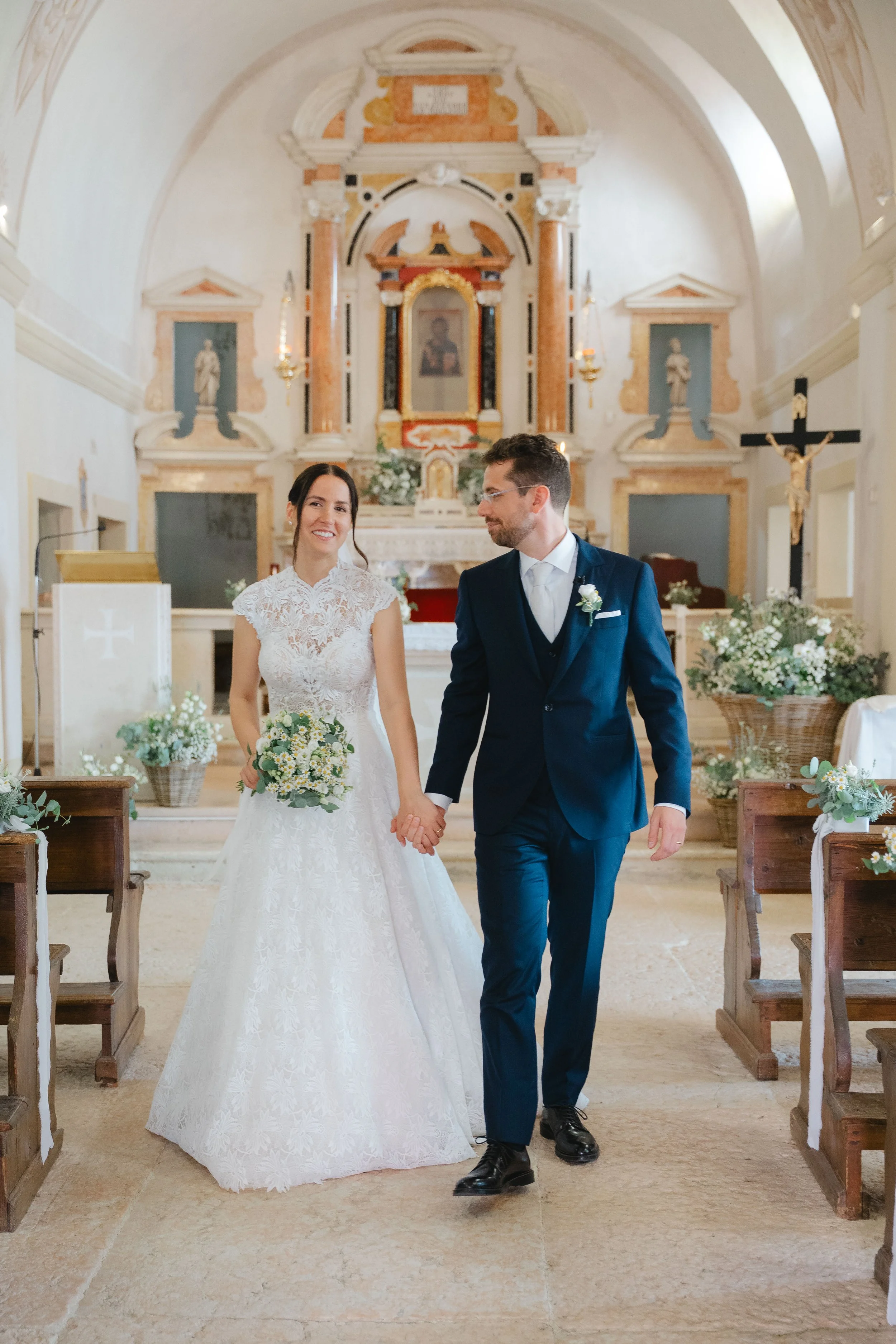 A bride and groom walking hand in hand down the aisle of a church, smiling, with floral arrangements on either side and religious icons and a large crucifix in the background.