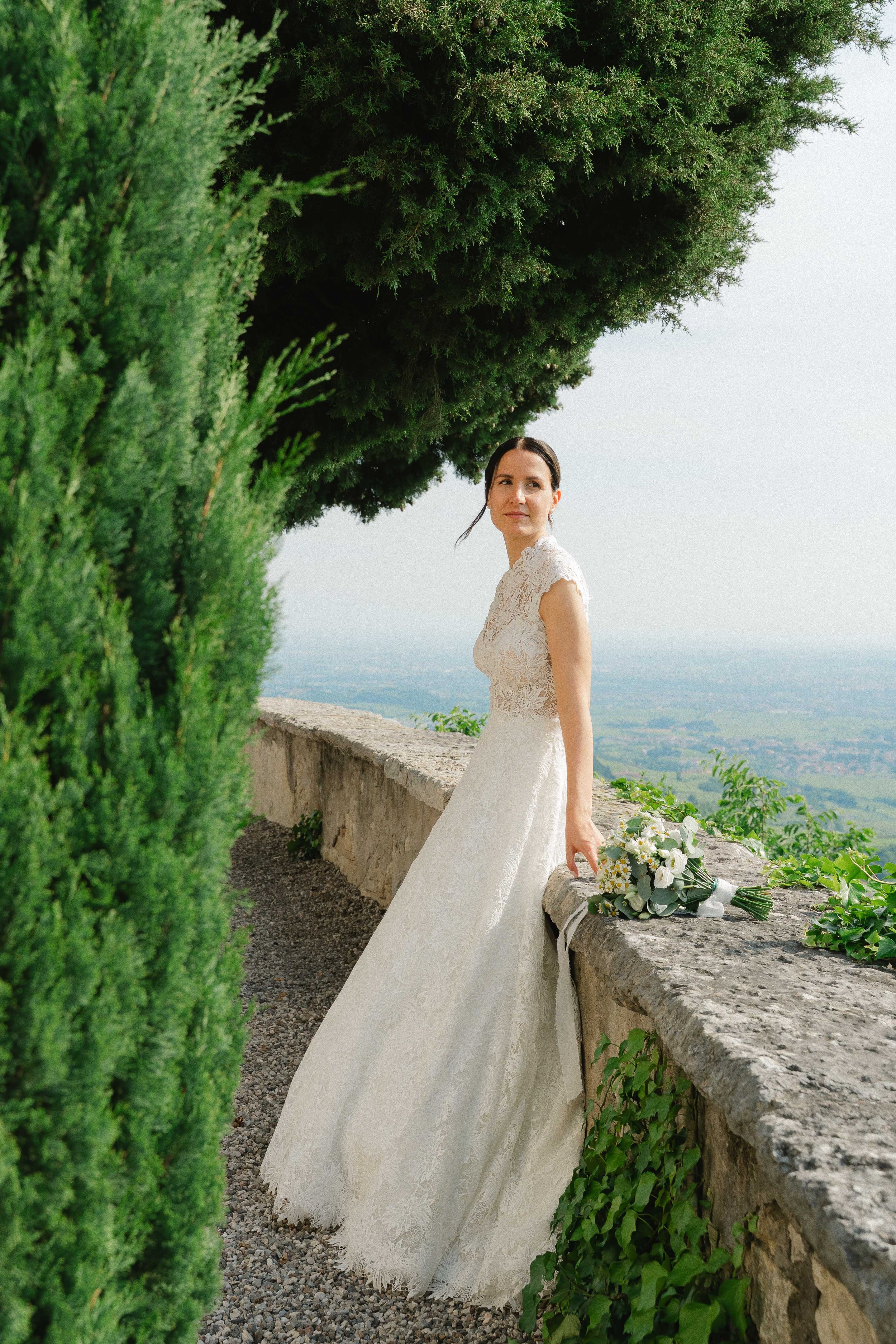 A bride in a white lace wedding dress stands beside a stone wall with a bouquet of flowers, overlooking a scenic landscape with green trees and a cloudy sky.
