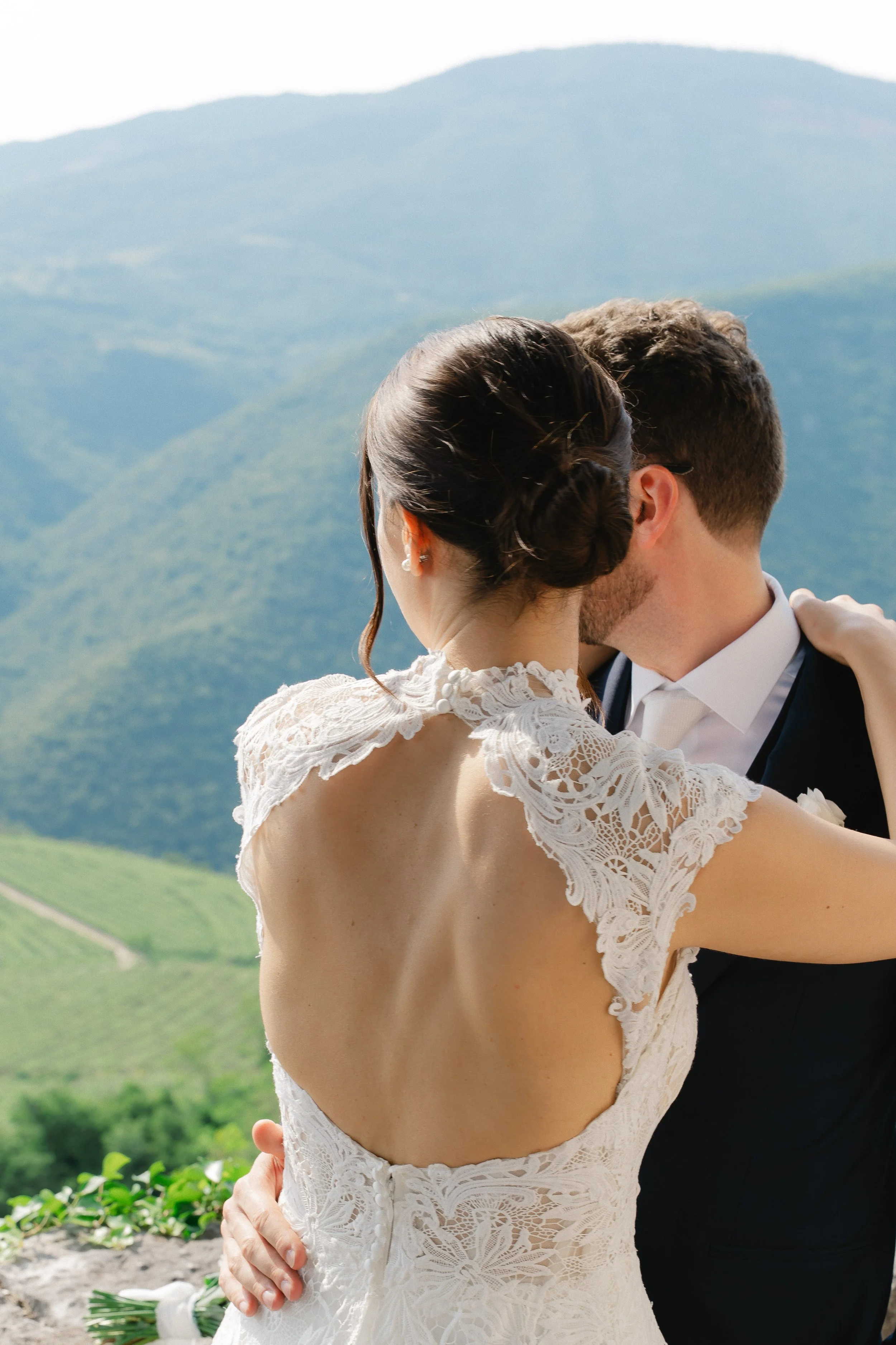 A bride and groom are embracing outdoors with a mountainous landscape in the background. The bride is wearing a lace wedding dress and has her hair styled in an elegant updo. The groom is in a dark suit and white shirt. The bride's bouquet is visible on the ground.