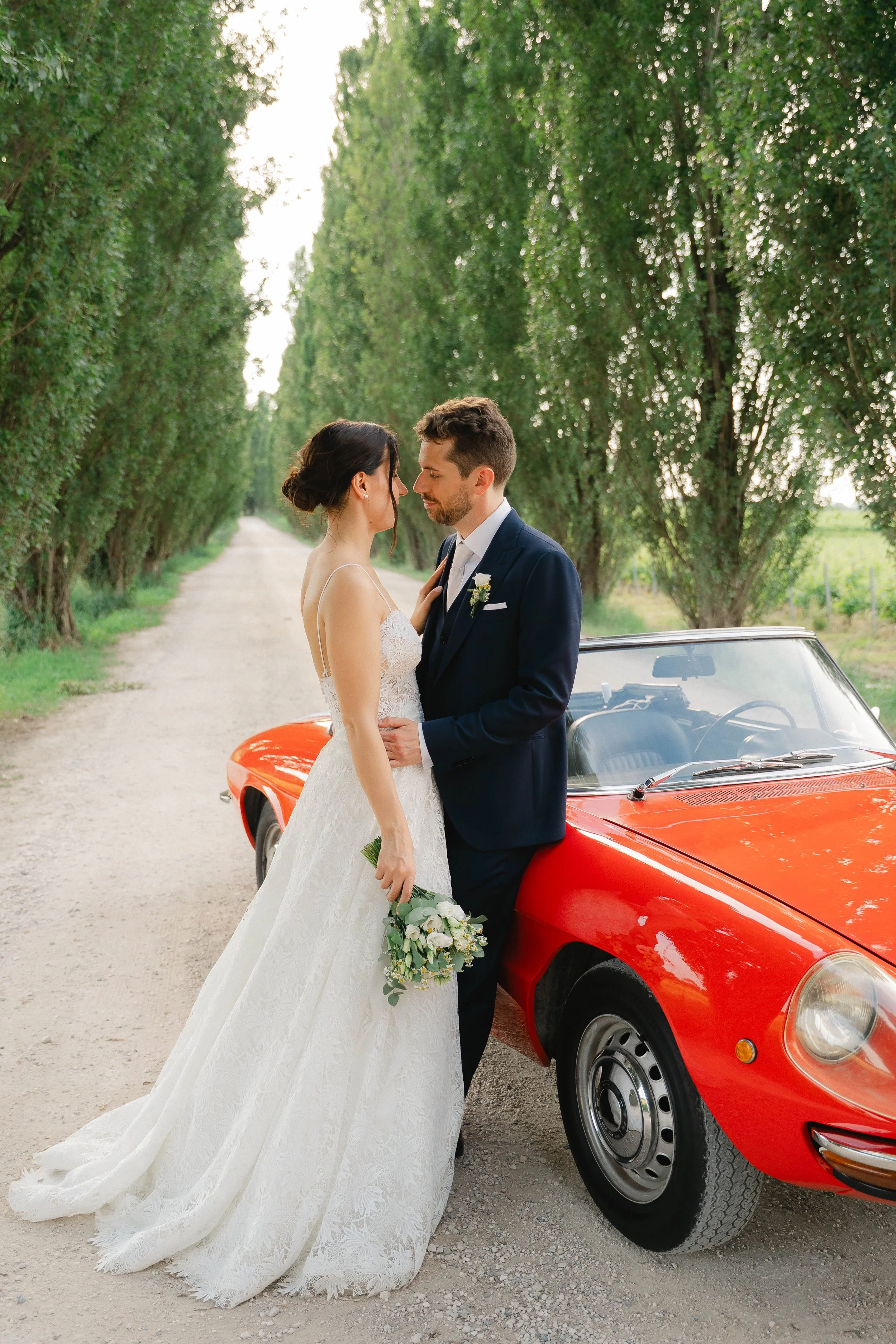 A bride and groom standing close together in front of a red sports car, gazing at each other, on a rural dirt road lined with green trees.