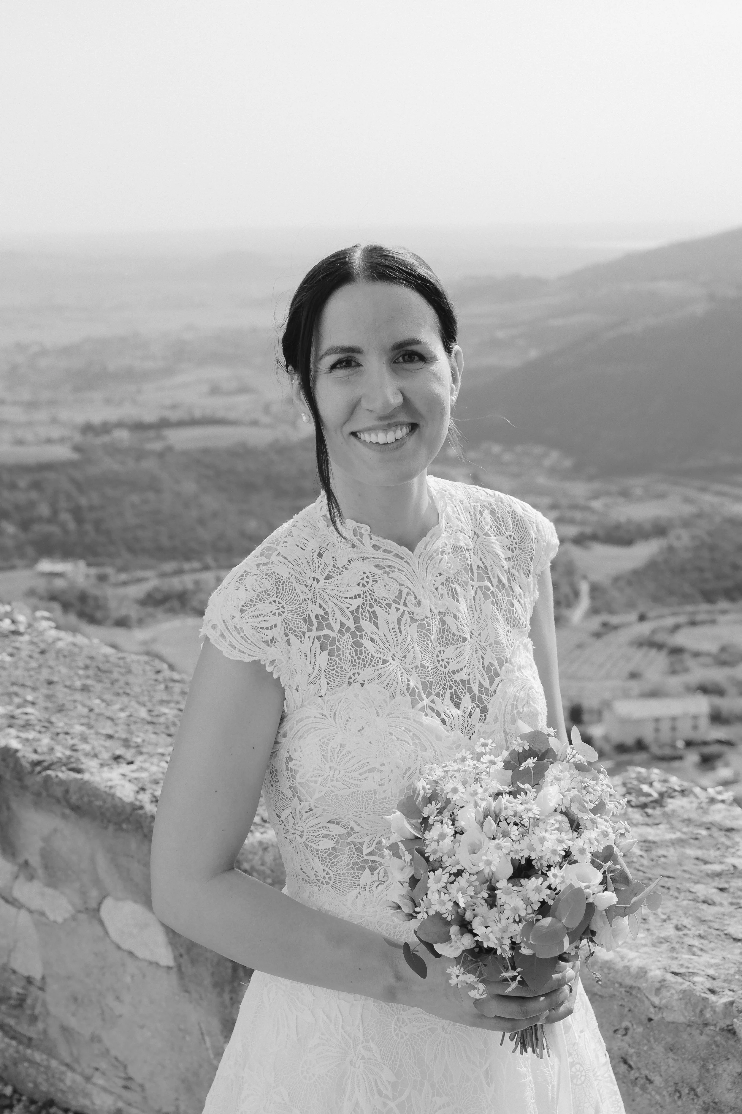 A woman in a lace dress smiling while holding a bouquet of flowers, with a landscape of hills and buildings in the background.