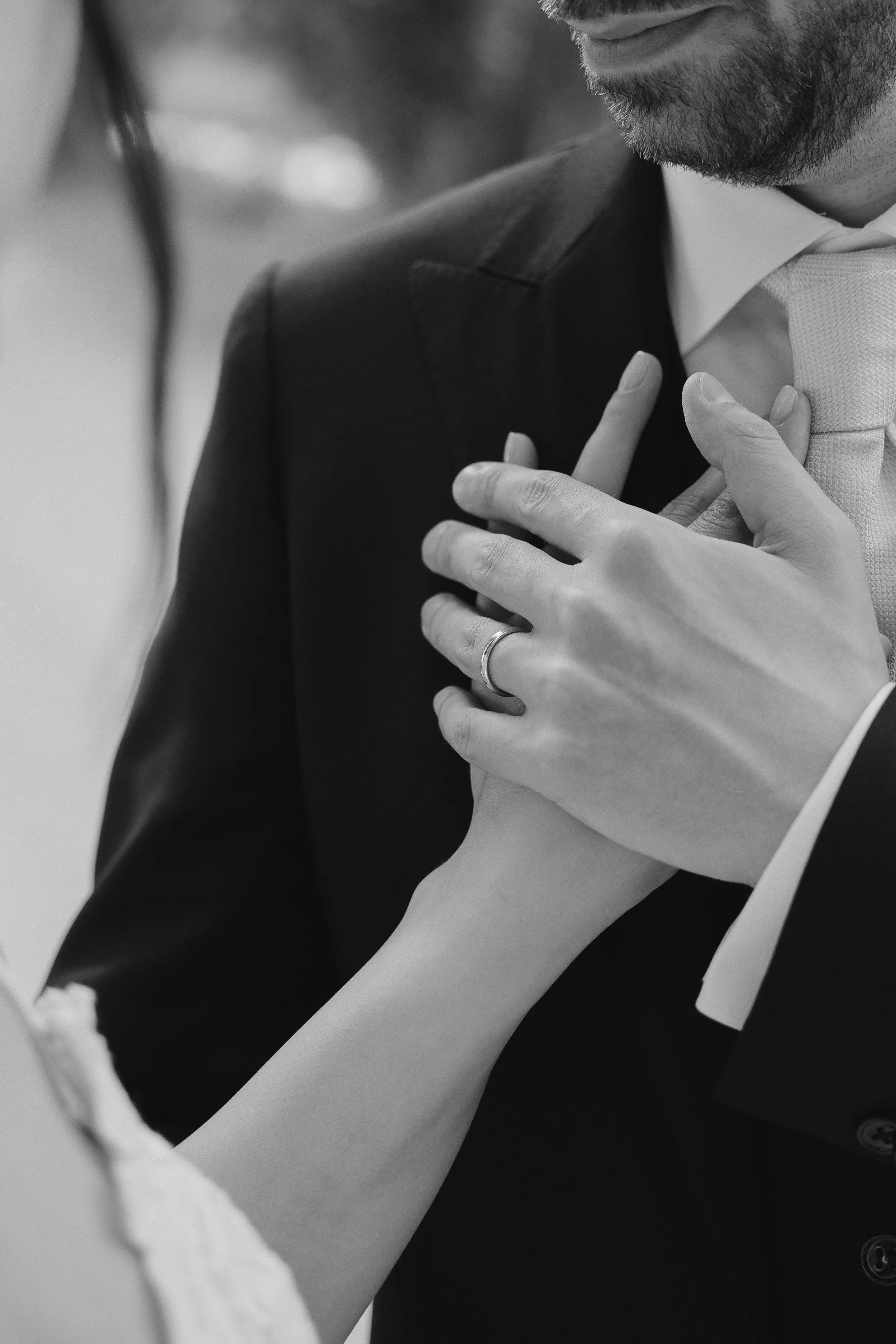 A black and white close-up photograph of a man in a suit with a beard, wearing a wedding band, gently holding the shoulder of a woman wearing a white dress or gown.