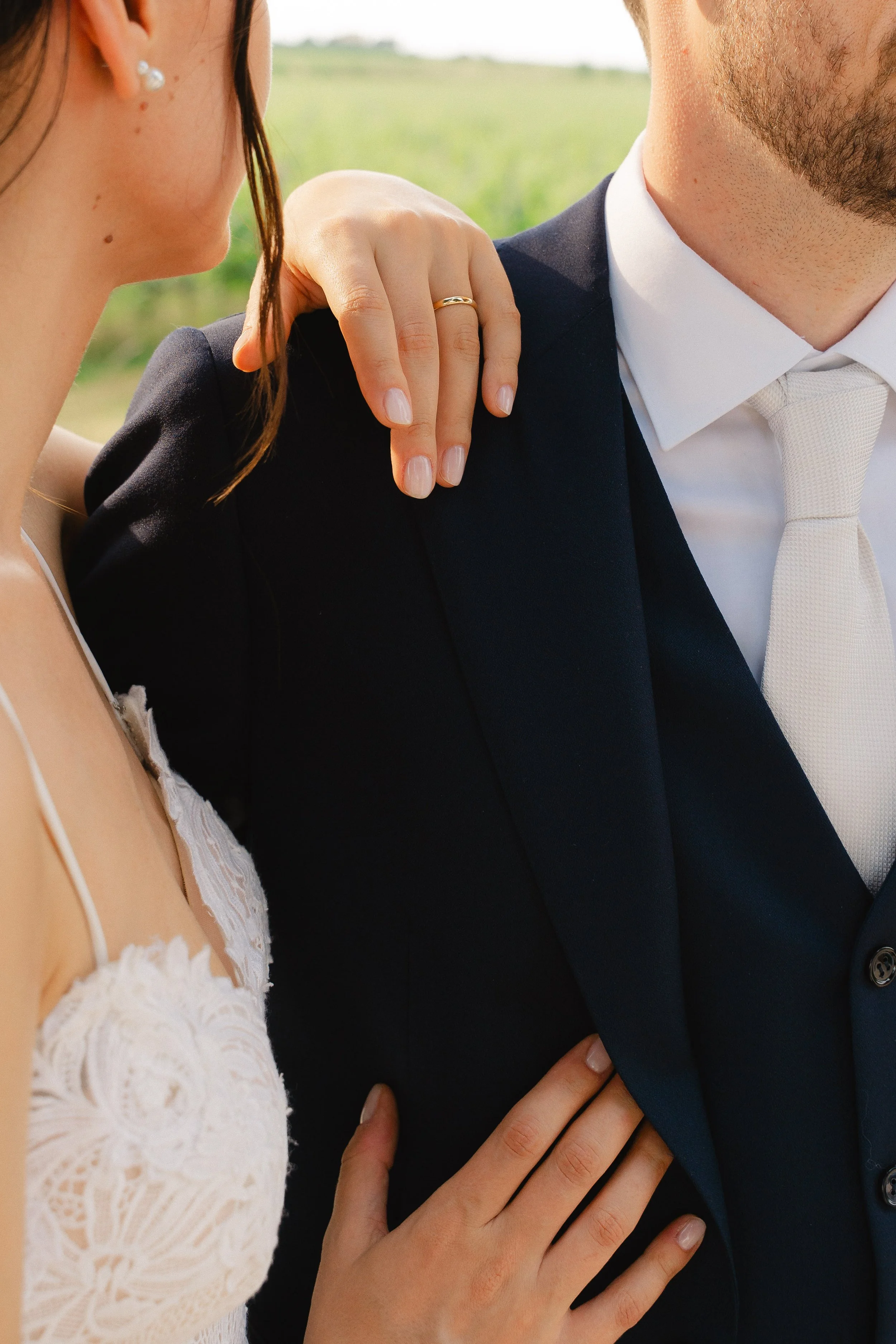 Close-up of a woman and man, focusing on their shoulders and hands. The woman is wearing a white lace dress and a pearl earring, and has a wedding ring on her finger. The man is dressed in a dark suit with a white shirt and a light-colored tie.