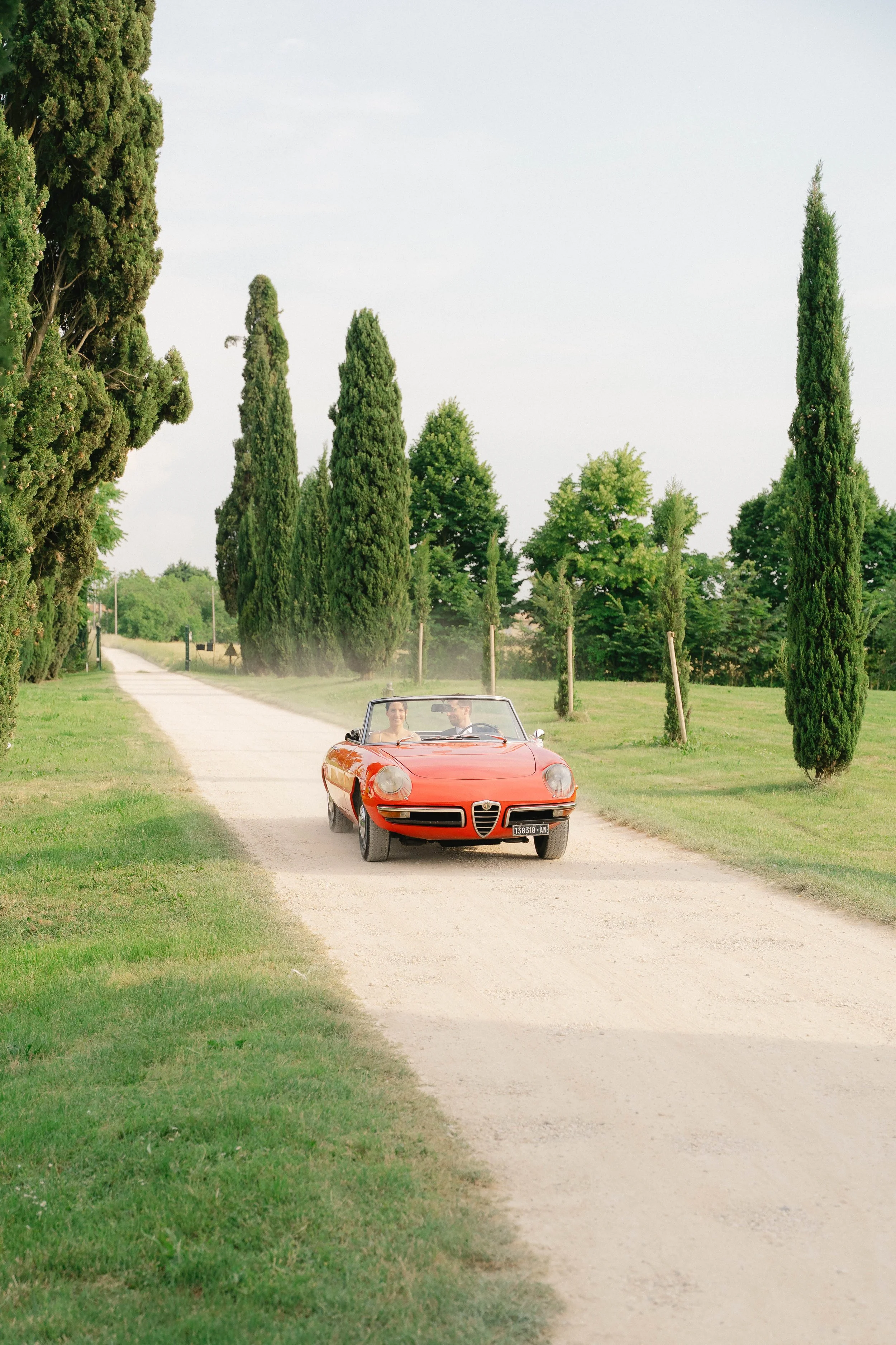 A vintage red convertible sports car driving on a gravel country road lined with tall green cypress trees, with two people inside smiling at each other.