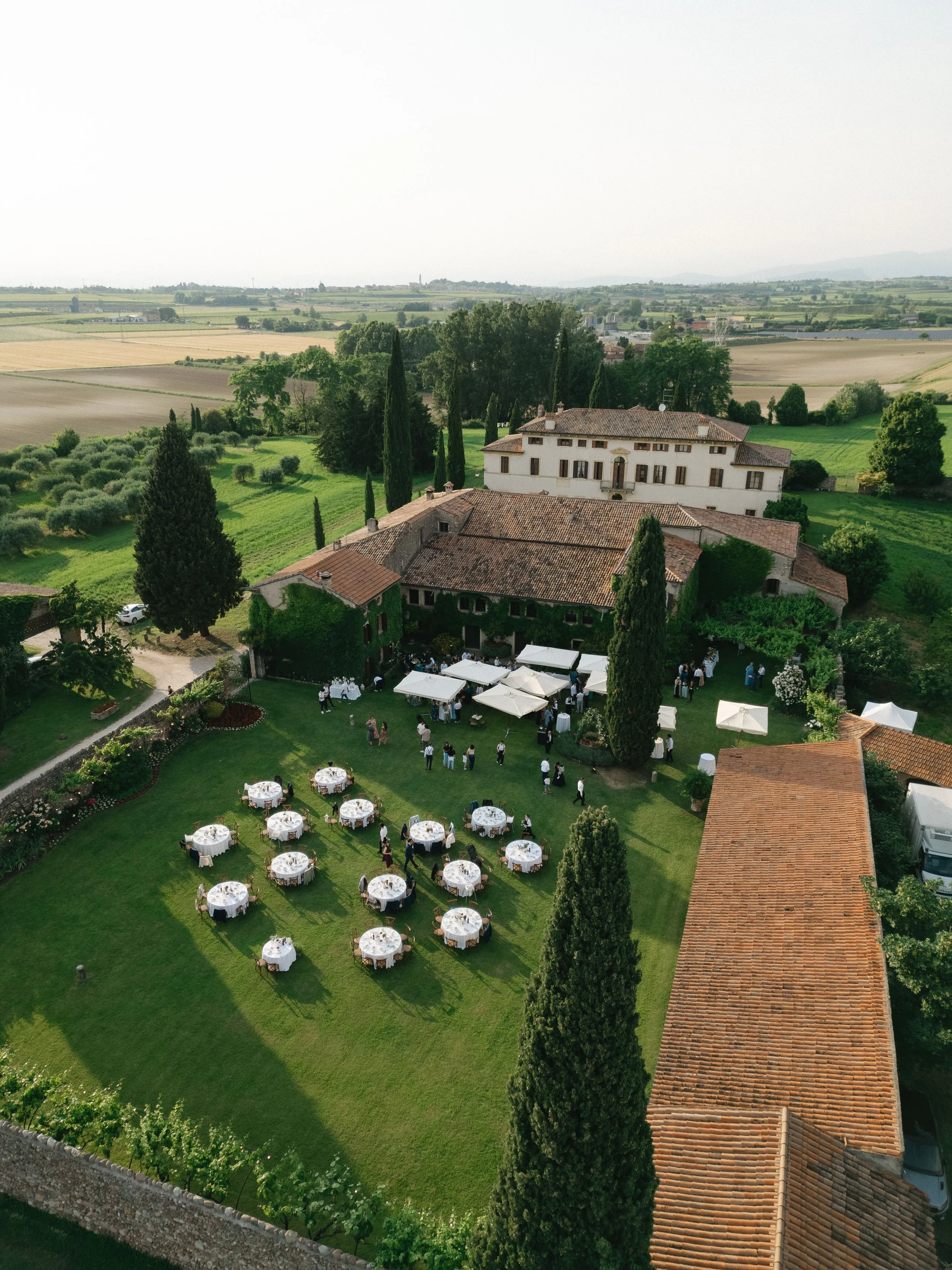 An aerial view of a countryside estate with a large white building, surrounded by trees and green lawns. Several round tables with white tablecloths and umbrellas are set up on the lawn, indicating a outdoor event or gathering. Rolling farmland extends into the distance on a clear day.