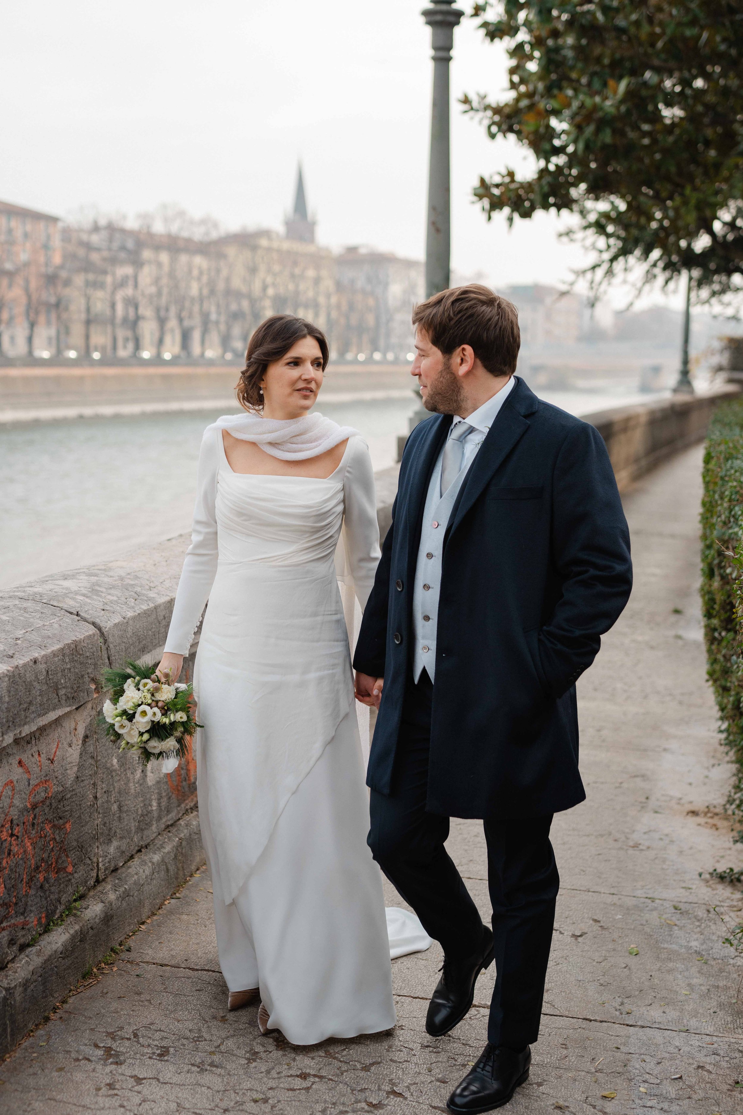 A bride and groom walking along a riverside pathway holding hands, with the bride holding a bouquet and wearing a white wedding dress layered with a white shawl, and the groom wearing a dark suit with a vest and tie, against a backdrop of leafless trees, urban buildings, and a church steeple.