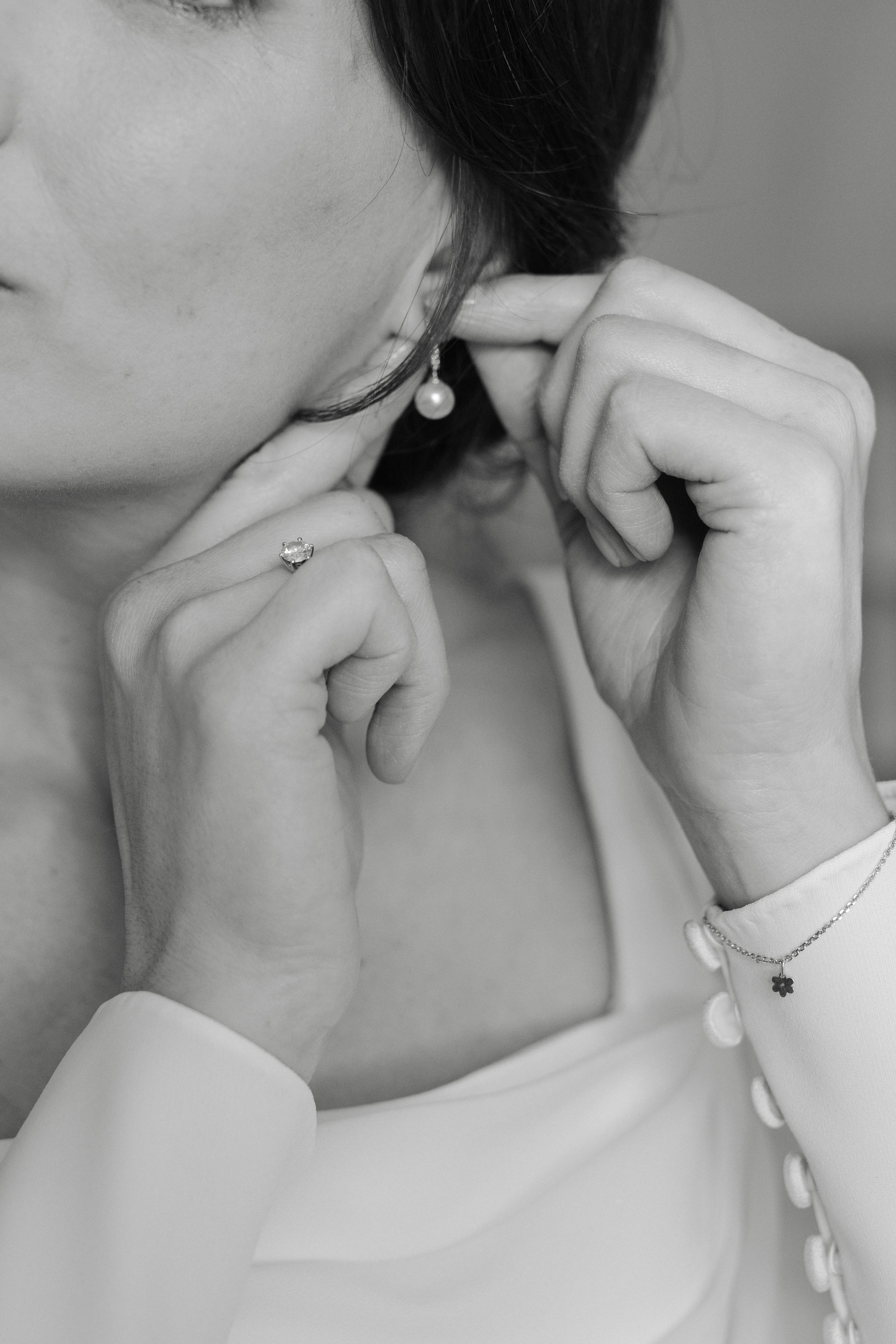 Close-up of a woman wearing pearl earrings, a ring, a necklace with a four-leaf clover pendant, and a buttoned blouse. She is touching her ear and neck with her hands and has dark hair.
